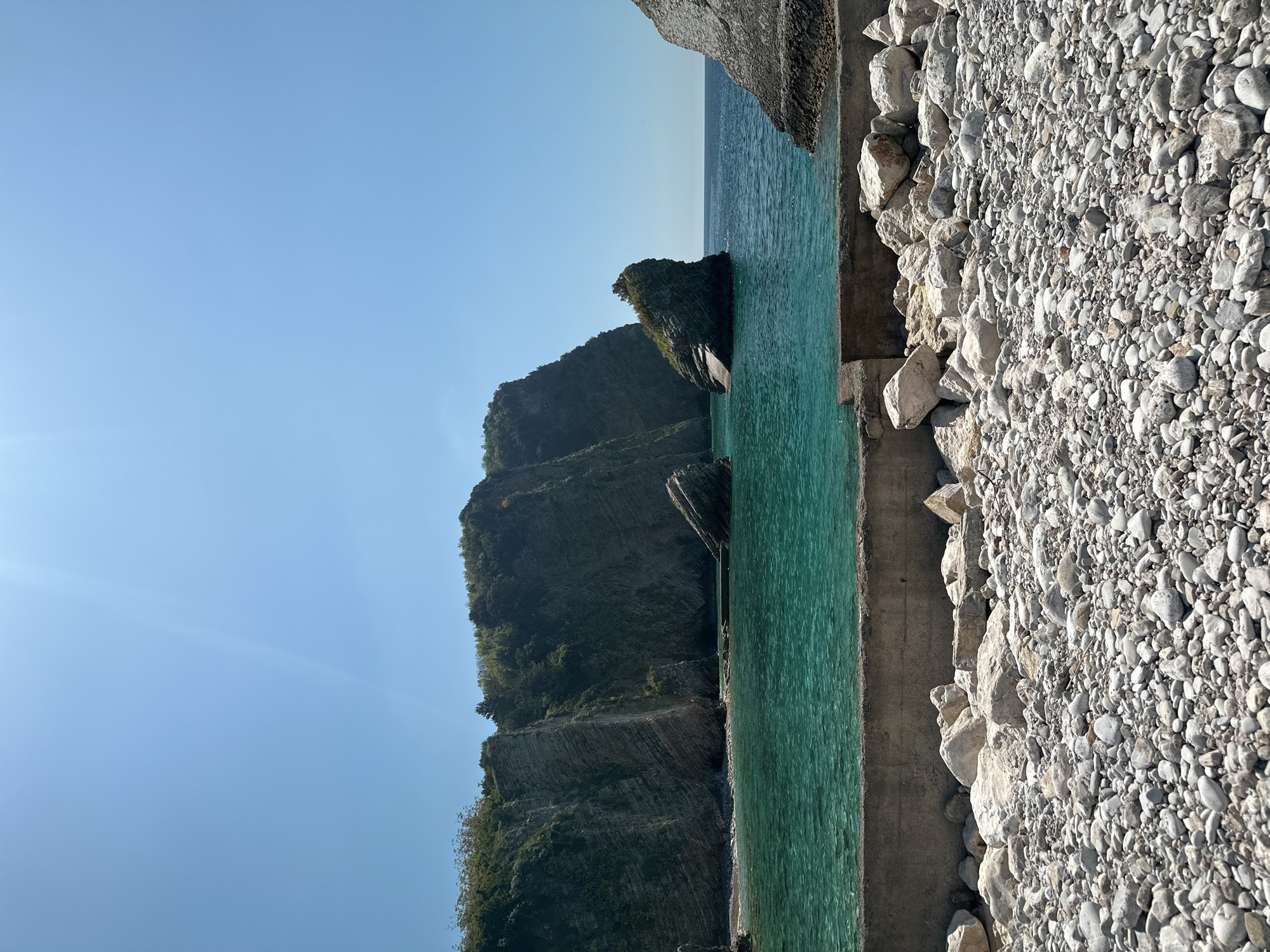 The scene depicts a coastline on a clear day, likely in Budva, Montenegro. The image is oriented vertically, showing the sky on the left, followed by a rock cliff dropping into the turquoise water. A few rock formations are situated within the water just off the cliff. On the right of the water's edge is a concrete seawall separating the water from a rocky beach composed of small, light grey pebbles and larger stones. No people are visible in the image.
