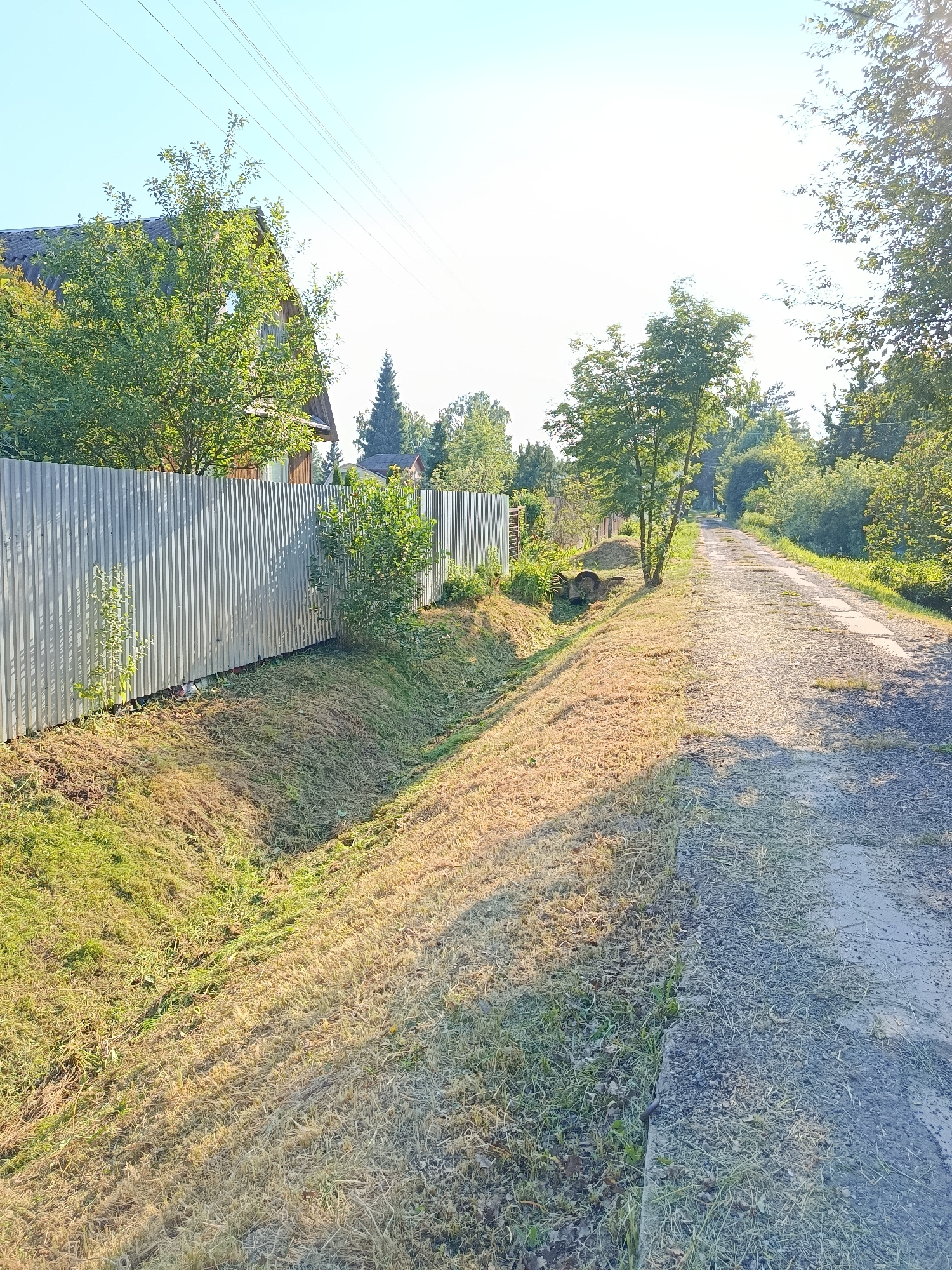 The image depicts a rural road in Alferovo, Russia, on a sunny day. The road is unpaved and appears to be a dirt track or gravel path. To the left of the road, there is a ditch with dry grass and some green vegetation. Beyond the ditch, a corrugated metal fence runs along the property line, separating the road from a residential area. A house with a wooden exterior and a dark roof is partially visible behind the fence, surrounded by trees and greenery. The sky is clear and blue, with a few power lines visible in the distance. The overall scene suggests a quiet, rural setting.