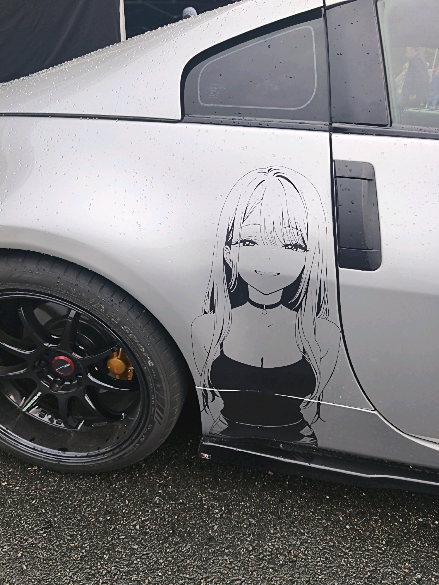 A silver car, possibly a Nissan 350Z based on the body style and wheel design, is parked on a wet gravel surface, suggesting an outdoor environment like a parking lot or roadside. The car's body is covered in raindrops, indicating recent or ongoing precipitation. A prominent black vinyl sticker of an anime-style female character with long hair, wearing a tank top and a choker, is affixed to the rear passenger door. The character has large eyes and a smiling expression. The car's black alloy wheel with a red accent and gold brake caliper is visible in the foreground. In the background, parts of a dark tent or canopy and a reflection of a person in a suit can be seen, possibly indicating an event or gathering. There is no discernible indication of the location being Tallinn, Estonia; the provided description is based solely on the visual content of the image.