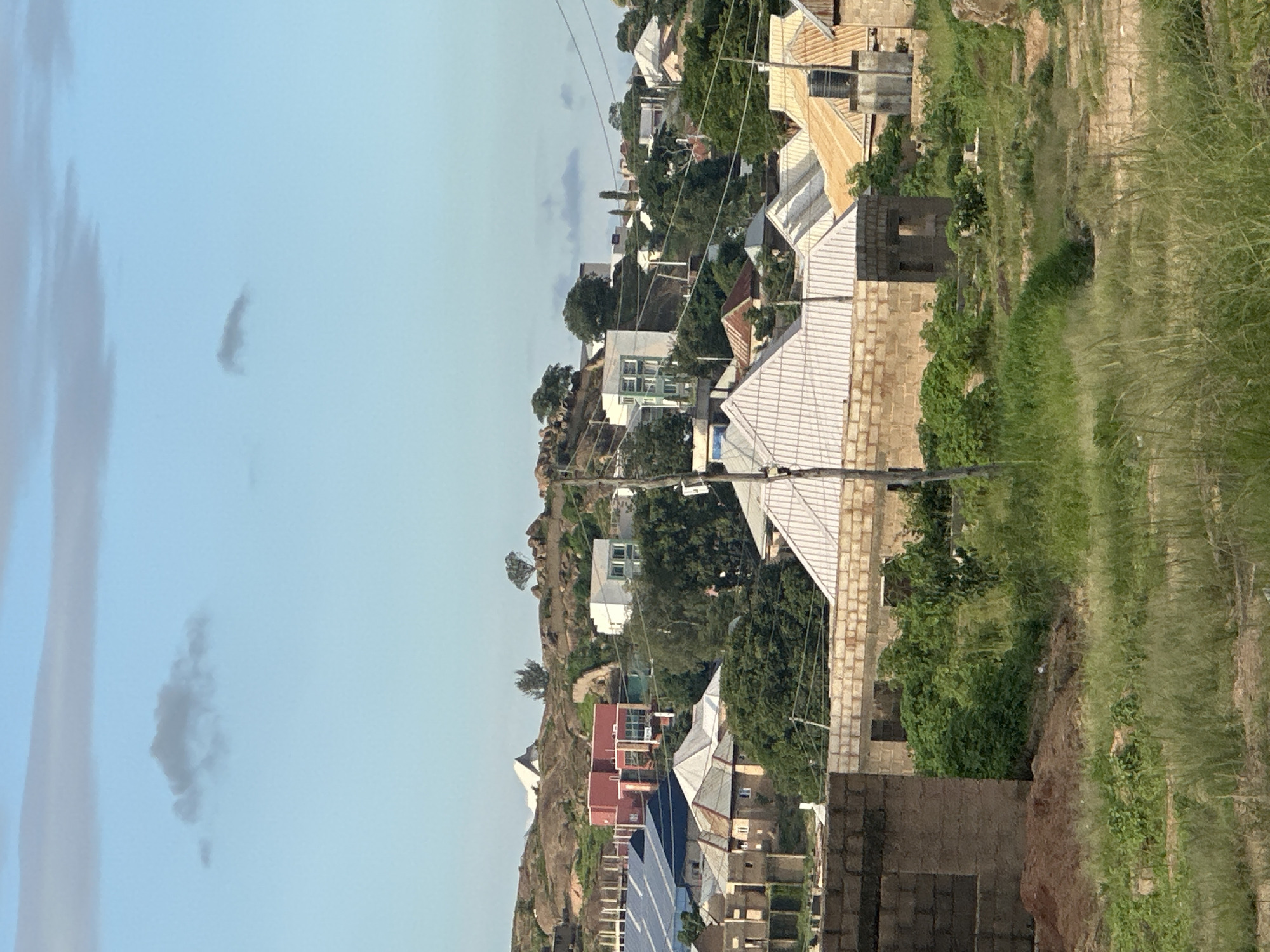 The image depicts a residential area on a hillside in Jos, Nigeria. The foreground shows an unfinished brick building and overgrown grass, suggesting ongoing construction or a neglected area. The midground is dominated by a variety of houses with corrugated metal roofs, some appearing newer and more modern, while others are older. A larger, red building with numerous windows stands out on the hillside. Further up the slope, more buildings are visible, interspersed with trees and rocky outcrops. The sky above is a pale blue with a few wispy clouds. The overall impression is of a developing urban landscape in a hilly terrain.