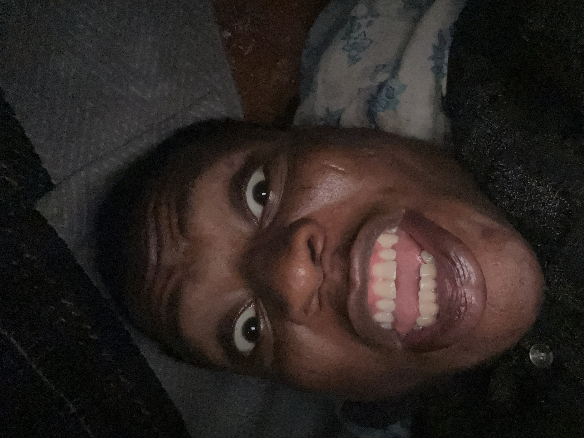 A close-up, high-angle portrait of a Black person with dark skin and short, dark hair. They are smiling widely, revealing a full set of white teeth. Their eyes are open and appear expressive. The lighting is dim, and the background is indistinct but suggests fabrics or bedding. The environment is undiscernible, and no location information is available.