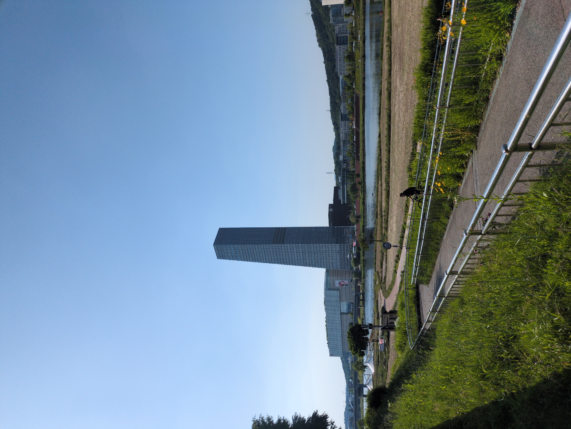 The image depicts a daytime scene in Daejeon, South Korea, featuring the Daejeon Government Complex Building 1. A paved pathway with metal railings runs along a grassy verge to the right, with a cyclist visible further along the path. A river or waterway is located beyond the pathway, separating it from buildings on the opposite bank. The sky is clear and blue.