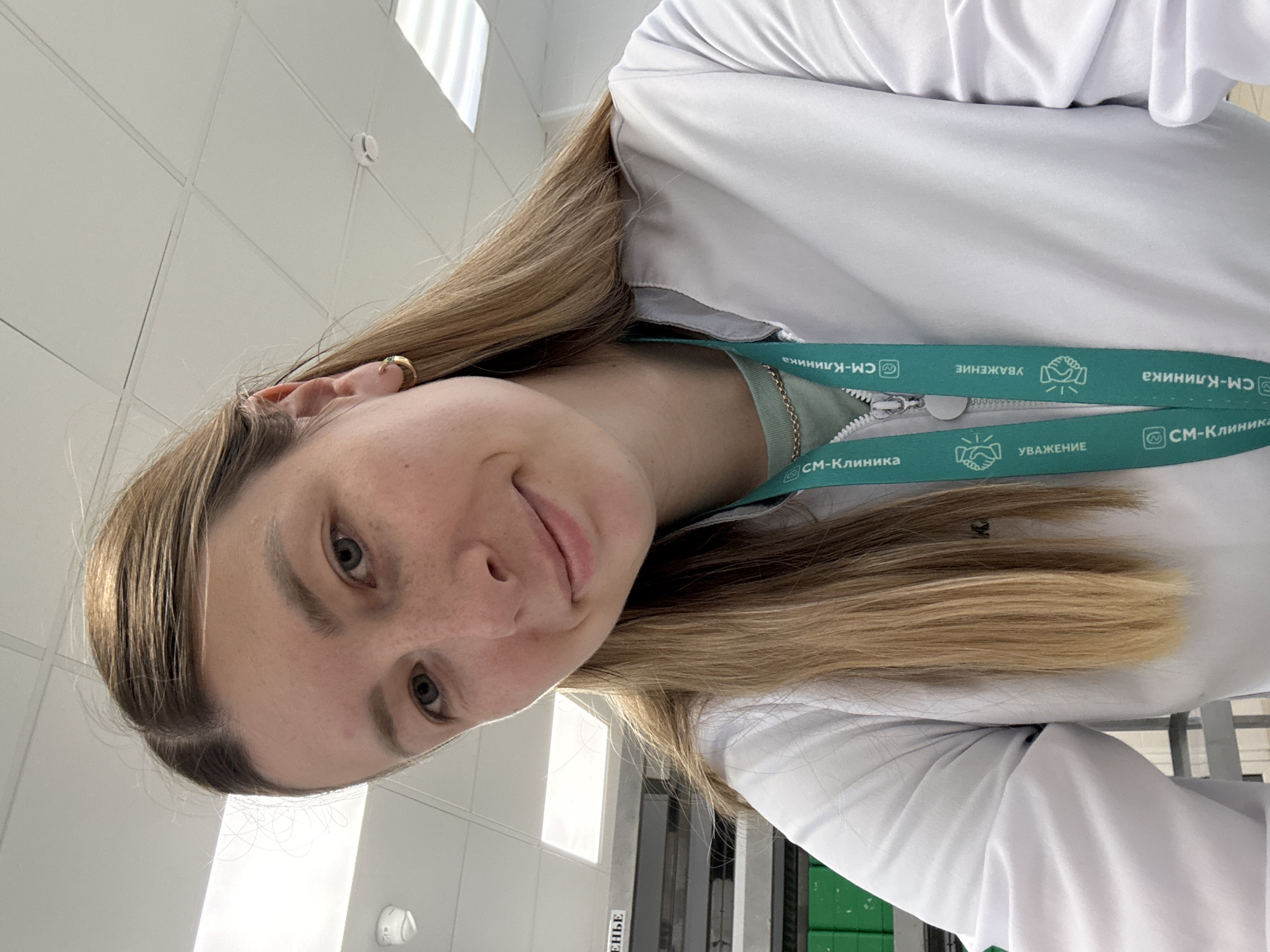 A woman with long blonde hair, wearing a white lab coat and a lanyard with the words "CM-Clinic" and "Respect" in Russian, is pictured indoors. The background includes a white tiled ceiling, a window, and a door. "CM-Clinic" suggests the location is likely related to a medical clinic. The Russian language on the lanyard indicates the location is in Russia, and based on the clinic's name, potentially Moscow.