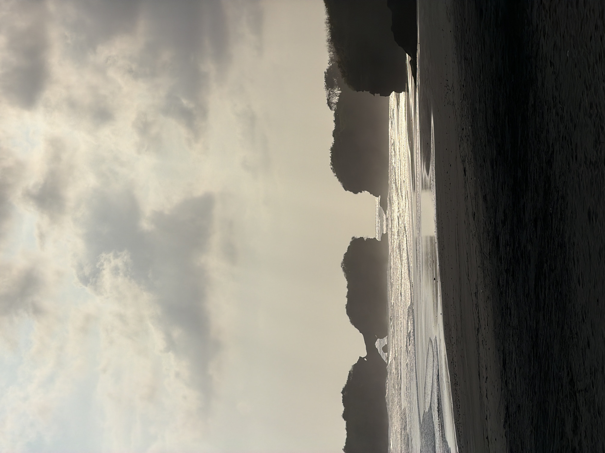 The image depicts a serene beach scene at Selemadeg, Indonesia, likely during either sunrise or sunset, given the soft, diffused light. The foreground is dominated by a dark, sandy beach. Beyond the sand, the ocean stretches out, with gentle waves lapping the shore and reflecting the muted sky.
Several prominent rock formations rise from the water, silhouetted against the bright horizon. The largest of these formations, located in the mid-ground, features an archway carved by the sea, connecting two massive rock pillars. These rocky outcrops are covered in vegetation, adding a touch of green to the otherwise monochromatic scene.
The sky is filled with a layer of soft, gray clouds, with some patches allowing the sun's light to filter through, creating a hazy, ethereal glow. The overall atmosphere is one of tranquility and natural beauty, capturing the essence of a quiet coastal landscape. There are no people or animals visible in the image, emphasizing the solitary and untouched nature of the location. The scene is captured in a vertical orientation, further highlighting the vastness of the sky and the imposing presence of the rock formations.