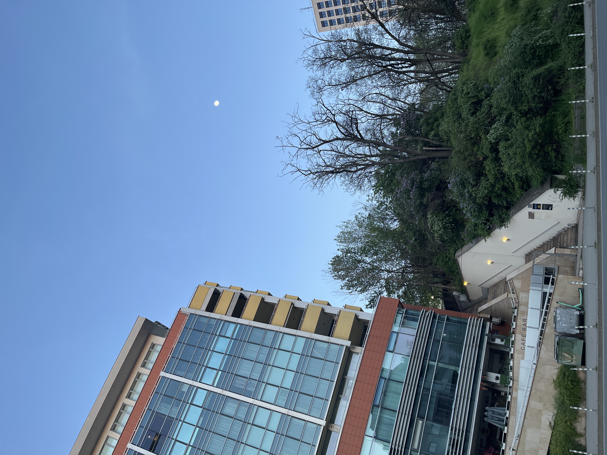 This image depicts an urban setting featuring modern architecture juxtaposed with a natural, green hillside, all under a clear blue sky.
In the foreground and to the left, a contemporary building dominates, characterized by large glass window panes reflecting the sky, and reddish-brown exterior panels. Its upper floors feature distinctive yellow, box-like balconies or architectural details.
To the right, a lush, green hillside rises, densely covered with trees and bushes. Some trees appear bare-branched, while others are in full foliage, with hints of purple flowers, possibly lilacs or jacaranda, visible amongst the greenery. A white building with a dark roof is nestled into the hillside, and a set of stairs winds upwards from the street level.
At the bottom right, a paved road or sidewalk is visible, lined with white and black bollards. An outdoor seating area with tables and umbrellas is situated next to the building. A sign clearly displays the text "CAFE BAR." A green electric scooter is parked near two trash bins (one grey, one green) by the roadside.
The sky is a vibrant, unblemished blue, indicating clear weather. A distinct, bright moon is visible in the upper left portion of the sky, suggesting either early morning or late afternoon/early evening. In the upper right background, another building with a grid of windows can be seen peeking over the trees. The image captures a blend of urban development and natural landscape, consistent with a city environment like Sofia, Bulgaria.