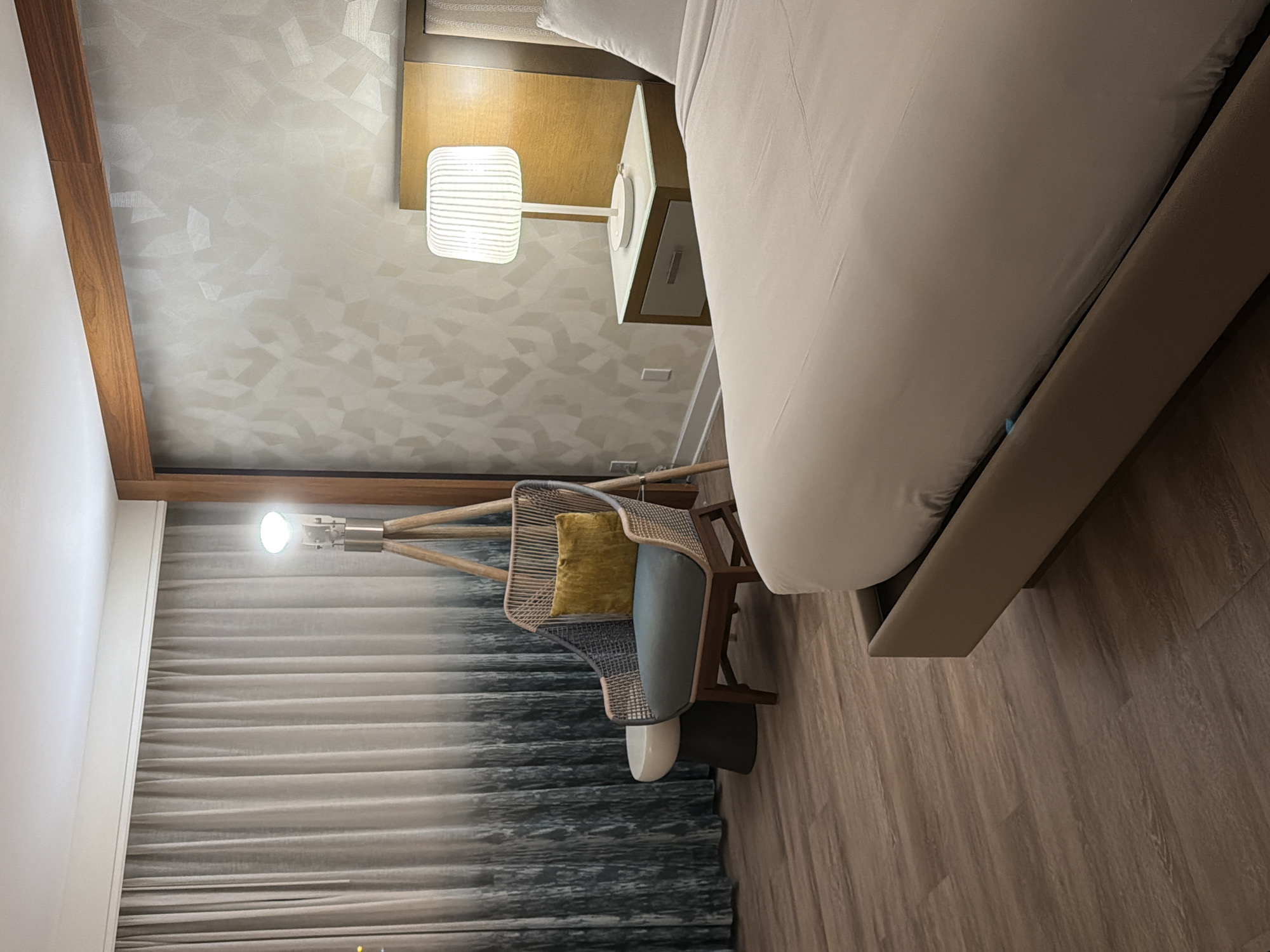 This is a photograph of a hotel room interior, likely in Newport Beach, California. The scene depicts a bedside area and a seating area with a floor lamp. In the foreground, a large bed with neutral-colored bedding is partially visible, with a sliver of the bed frame in tan upholstery. To the right of the bed is a nightstand with a grey drawer and a white lamp with a ribbed shade. Behind the nightstand is a headboard and part of a wall with a textured, geometric pattern in a light neutral tone. To the left of the bed, a rattan armchair with blue cushions and a yellow throw pillow is positioned next to a small, dark cylindrical stool. Behind the chair, sheer curtains with a gradient pattern, transitioning from white at the top to a darker, abstract pattern at the bottom, cover a window. A tripod floor lamp with a light bulb is situated behind the armchair. The flooring is made of wood-look planks. The lighting suggests it is nighttime or the room is dimly lit.
