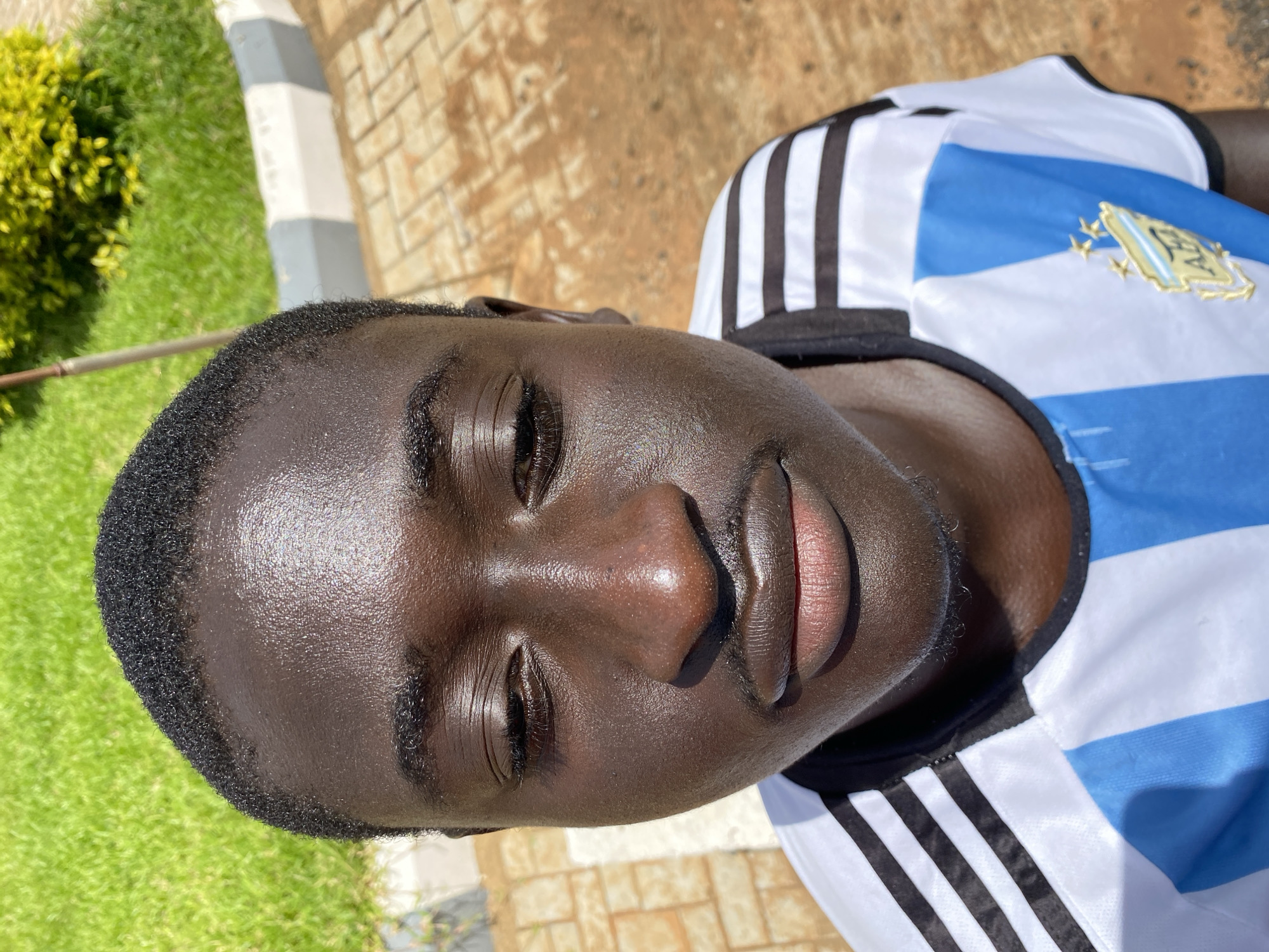 The image shows a close-up, portrait-style shot of a young Black person wearing a white and blue striped jersey, likely a soccer jersey, with the Argentinian crest visible on the chest. The person's face is tilted upwards and slightly to the right. Their skin is dark and appears to be glistening with sweat, suggesting warm weather. The background is blurred but shows hints of green grass, a paved area with interlocking bricks, and a raised border with gray and white stripes. The lighting indicates it is daytime, likely sunny, with shadows visible. The overall impression is of a casual, outdoor setting, possibly after physical activity. The person's expression is neutral. The image is taken from a slightly high angle, looking down at the subject.