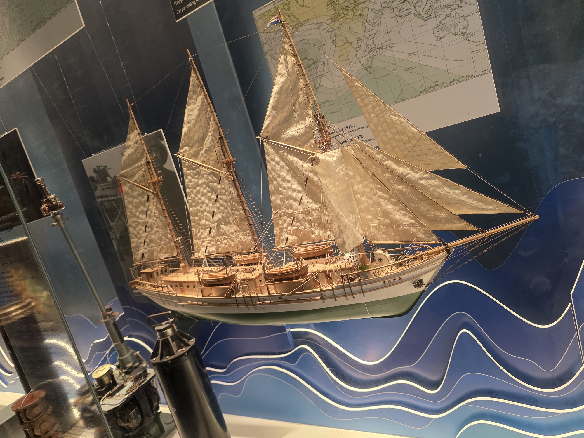 The image shows a detailed model of a sailing ship displayed in what appears to be a museum exhibit. The ship model is positioned at an angle, showcasing its numerous sails, rigging, and deck details, including lifeboats. Behind the ship, a world map is visible, suggesting a nautical or exploration theme. To the left of the ship, part of another exhibit is visible, including metallic scientific instruments enclosed in glass, likely related to maritime research. The background is a dark blue wall with stylized white waves, creating a watery backdrop. The lighting highlights the model ship and the instruments. The provided information indicates this is a museum exhibit in Kaliningrad, Russia, specifically within the Museum of the World Ocean, which houses extensive collections related to maritime history, exploration, and oceanography.