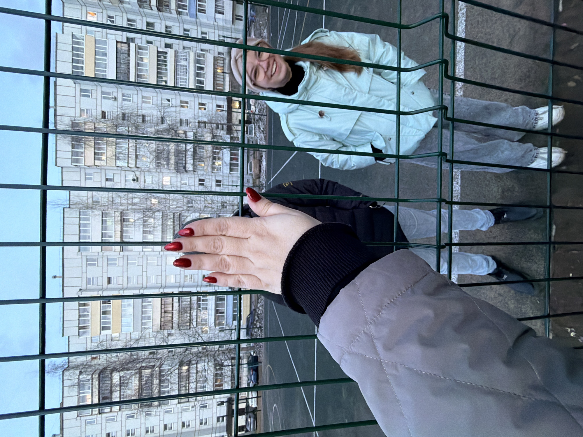 The image, taken through a green wire mesh fence, captures a moment between two individuals in an urban setting. In the immediate foreground, a person's right hand, adorned with bright red nail polish, is prominently featured, grasping or resting against the fence. This person wears a gray quilted jacket with a black knitted cuff.
Behind the fence, another person is visible, smiling widely and looking directly towards the camera. This individual has long, light brown hair, wears a light mint green puffer jacket, light-colored jeans, white sneakers, and light-rimmed glasses, topped with a light beige beanie. They are standing on what appears to be an outdoor sports court, evidenced by dark ground and faint white line markings.
In the background, several tall, multi-story residential apartment buildings dominate the skyline, featuring numerous windows and various balcony styles. Bare-branched trees are scattered between the fence and the buildings, suggesting a cooler climate or season. The sky is overcast, indicating either a cloudy day or dusk. The overall impression is a cheerful, casual interaction within a typical Russian residential area, specifically Chaikovsky, given the context. There is no visible text in the image.