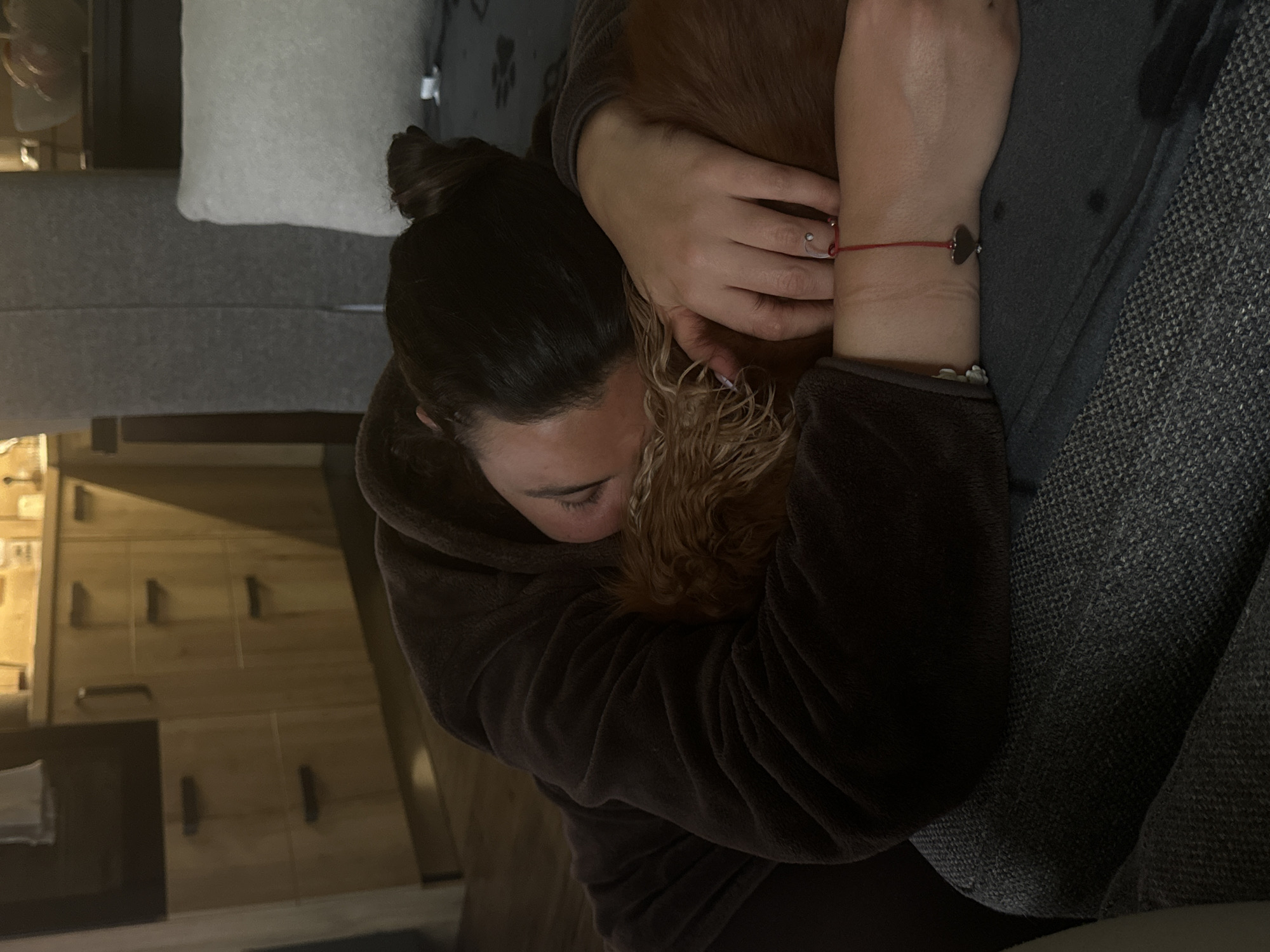 A woman in a brown robe is seated on a gray patterned couch and hugging a golden-brown dog. She has dark hair pulled into a bun. Her left wrist has a red string bracelet. The setting appears to be indoors, with wooden cabinets and drawers visible in the background. The location in Sofia, Bulgaria, cannot be determined from the image.