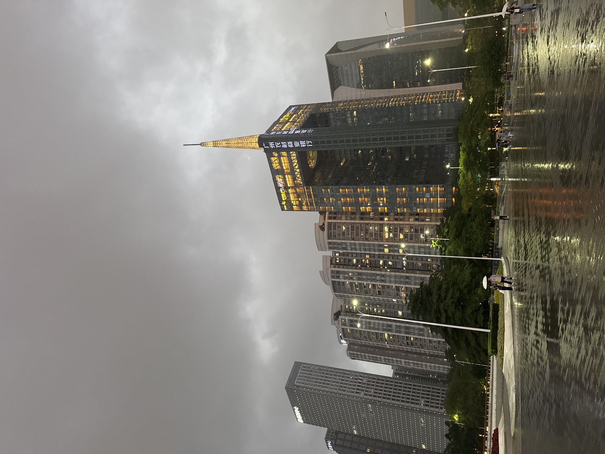 The image captures a cityscape in Tianhe District, Guangzhou, China, during a rainy evening. The foreground features a wet pavement reflecting the surrounding lights, creating a shimmering effect. A few people, some with umbrellas, walk along the path. Tall buildings dominate the background, with a prominent skyscraper distinguished by its gold spire and illuminated Chinese characters. Streetlights are sparsely visible along the sidewalk. The sky is overcast and grey.