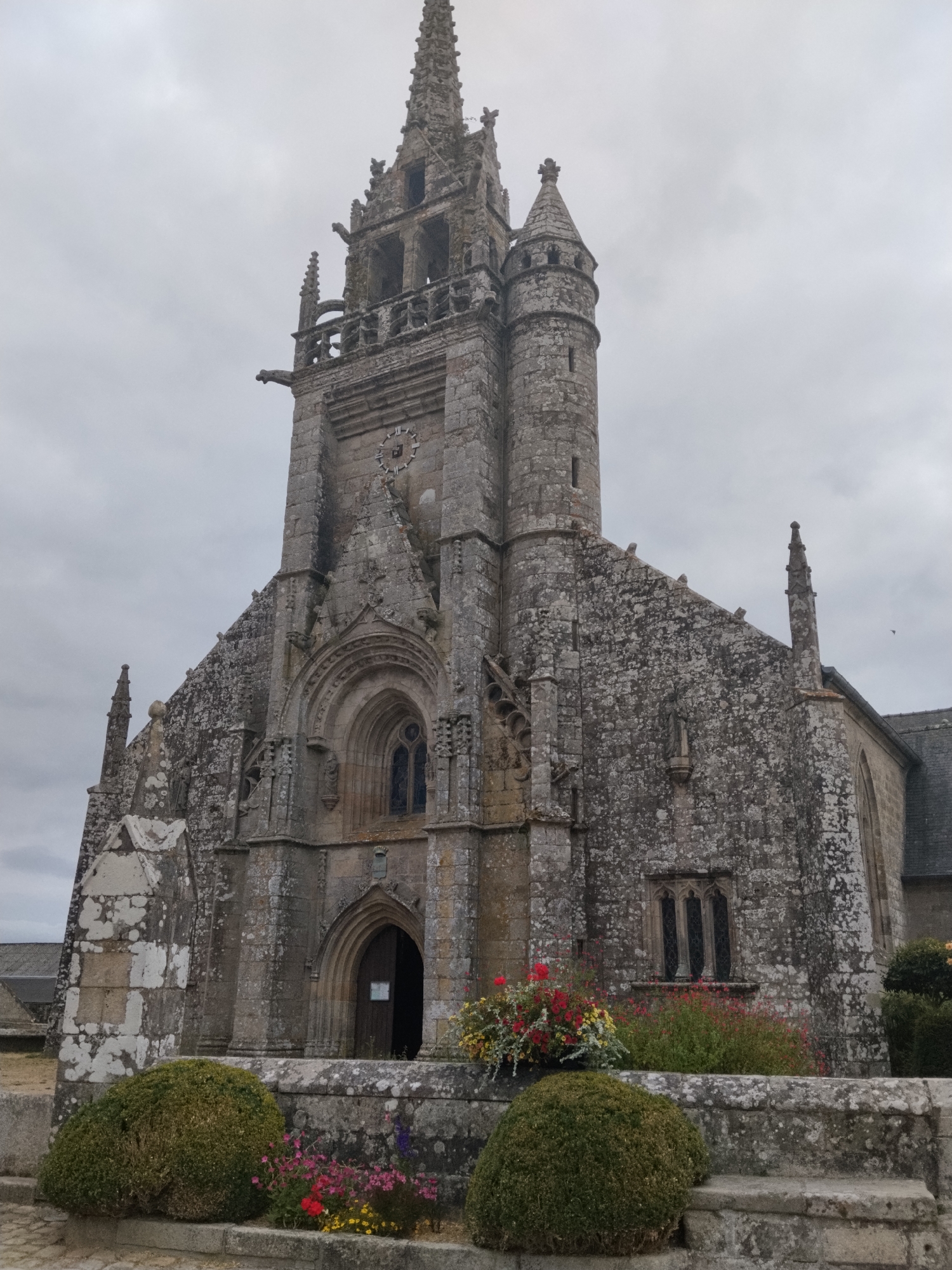 The image shows the exterior of a stone church, the Church of Saint-Pierre, in Guerlesquin, France. The church has a tall, intricate bell tower with a spire, gargoyles, and decorative stonework. A clock face is visible on the tower. The main facade features a large arched entrance with wooden doors and a gothic-style window above. Smaller windows are also visible on the church's walls. The building is weathered, with moss and lichen growing on the stone. In the foreground, there are two manicured green bushes and a stone wall with a flower planter filled with red, pink, and yellow flowers. The sky is overcast and cloudy. No people are visible in the image.
