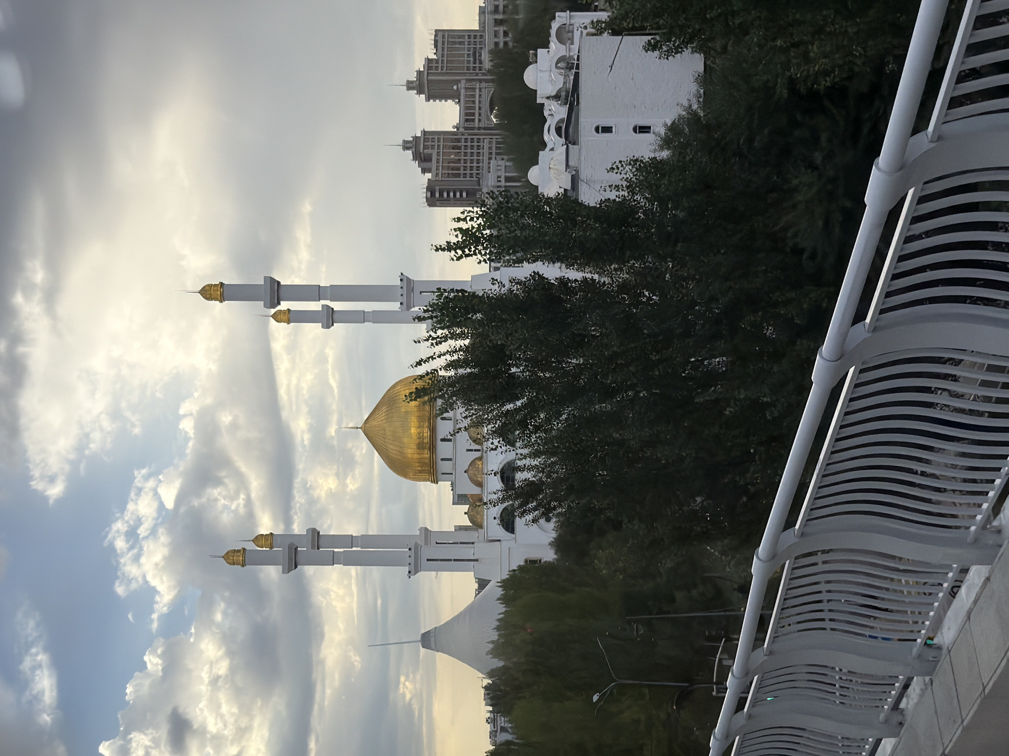 The image shows a view of the Hazret Sultan Mosque in Astana, Kazakhstan. The mosque features a large golden dome and two tall minarets with golden spires. The mosque is white and appears to be constructed from stone or concrete. In the background, other buildings, including a large white conical structure and several multi-story buildings, are visible. Dense green trees partially obscure the lower portions of the mosque and the surrounding structures. In the foreground, a white railing with vertical supports is visible, suggesting the photo was taken from an elevated position, possibly a bridge or walkway. The sky is partly cloudy with a soft, diffused light, indicating it may be early morning or late afternoon. No people are visible in the image.