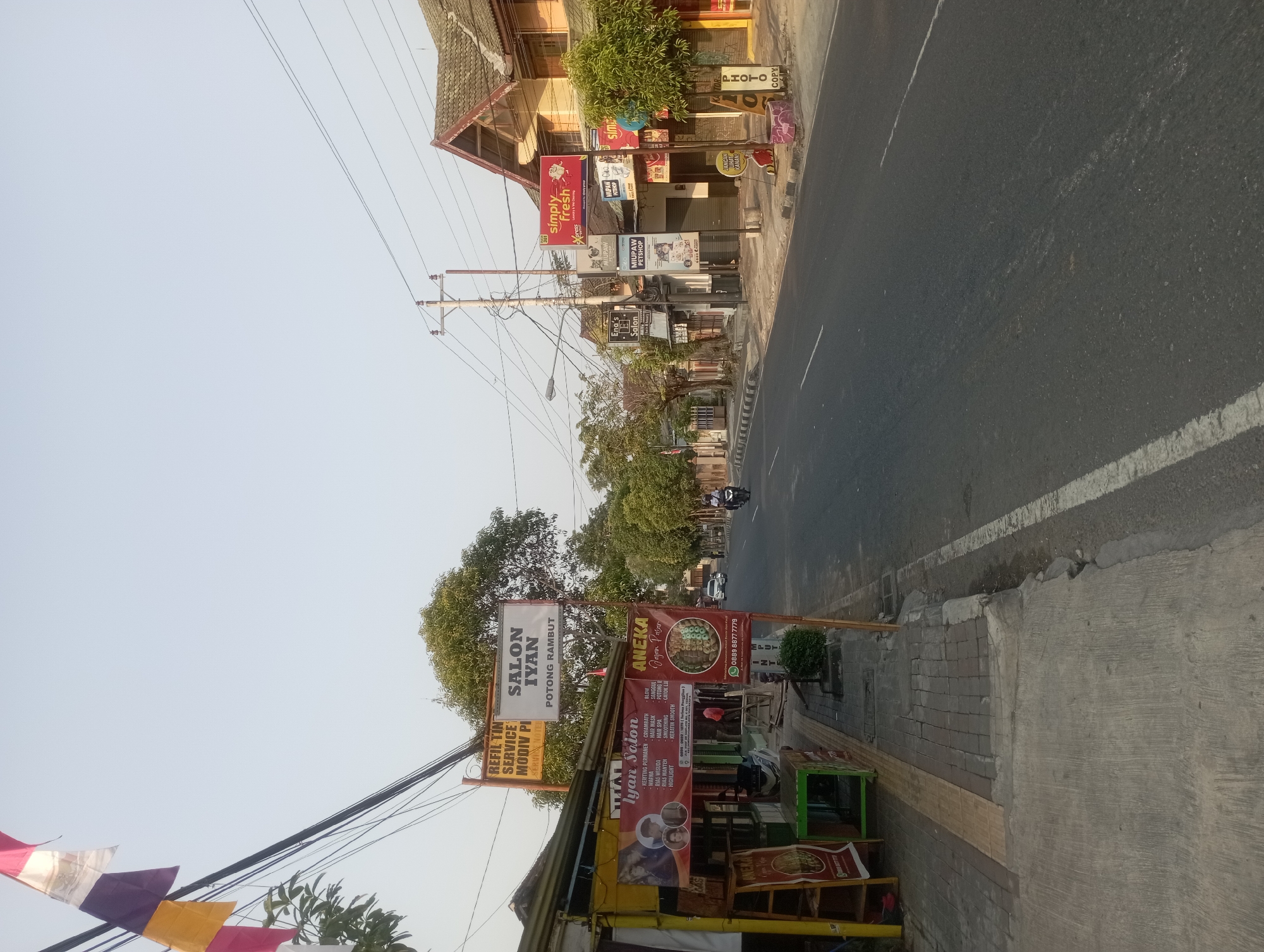 The image captures a street scene in Yogyakarta, Indonesia. On the left side of the road, a row of shops is visible. One shop displays a banner advertising "IYAN Salon" with a list of services including hair coloring, blow-drying, and smoothing. Above this, a sign for "SALON IYAN" with "POTONG RAMBUT" (haircut) written below it is prominently displayed. Another sign further up reads "REFIL TIN SERVICE MODIV PL".
Across the street, a multi-story building with a tiled roof is visible. Several shops line the ground floor of this building. Signs indicate a "simply fresh" laundry and dry cleaning service and a "MIUPAW PETSHOP". Further down the street, a motorcycle is being ridden by a person wearing a helmet, and a car is driving in the opposite direction.
The street itself is paved with asphalt and has white lane markings. The sidewalk is made of concrete and paving stones, with some areas showing wear and tear. Overhead, electrical wires are strung between utility poles. The sky is clear and bright, suggesting daytime. The overall impression is of a typical urban street scene in Indonesia, with a mix of businesses and daily life unfolding.