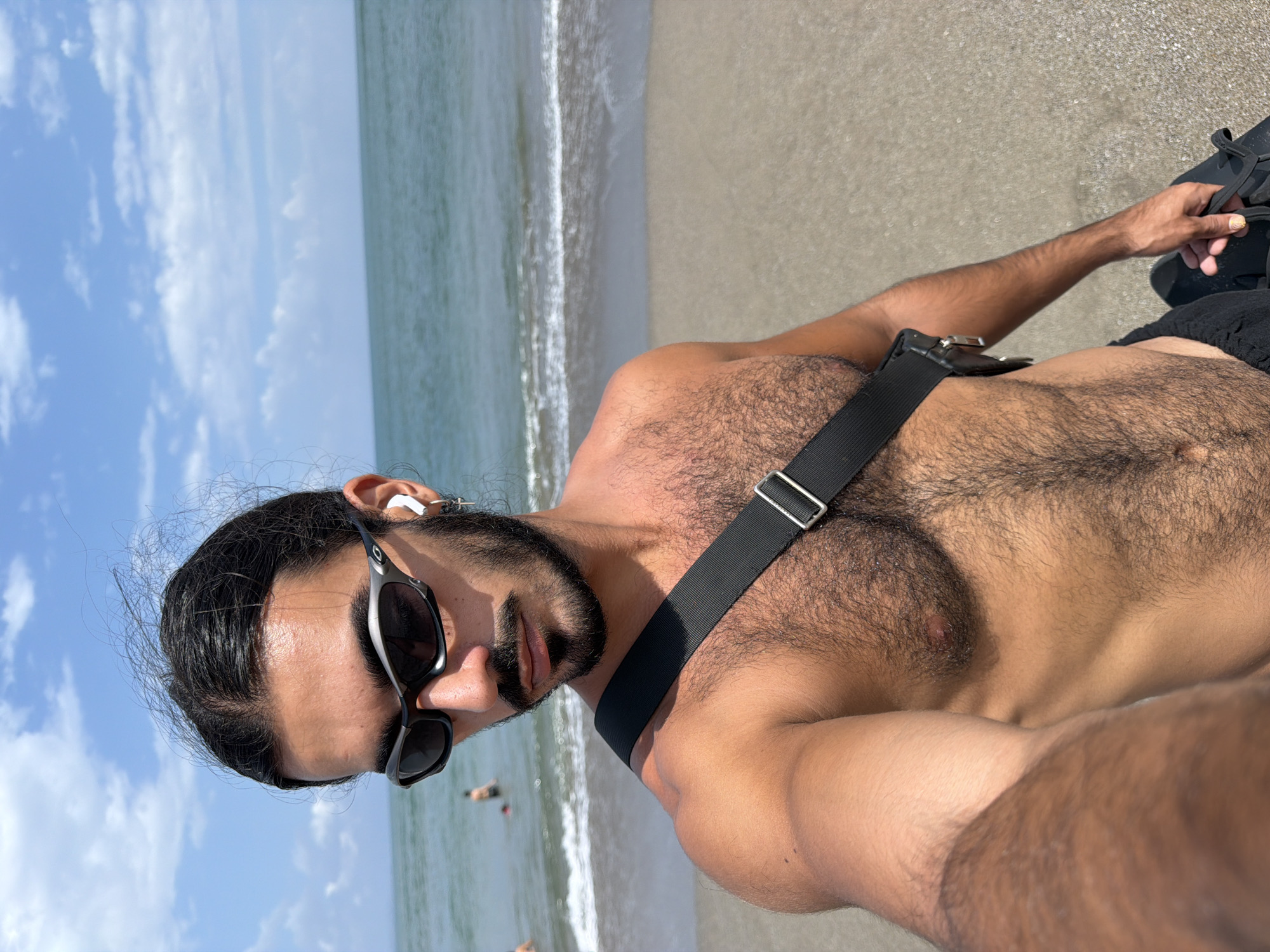 A shirtless man with dark hair pulled back in a bun, sunglasses, and white earbuds stands on a sandy beach. He has a dark beard and mustache and is wearing a black strap across his chest. The ocean is visible behind him, with gentle waves breaking on the shore. The sky is blue with scattered clouds. In the background, two figures are visible in the water. The man is holding a pair of black shoes in his right hand. This scene is consistent with a casual day at the beach in North Kuta, Indonesia.
