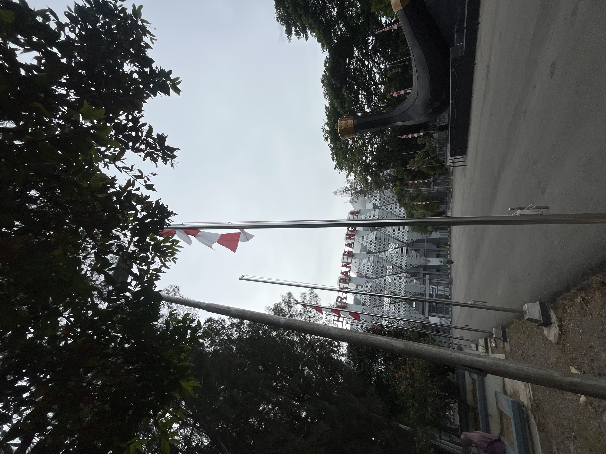 The image shows an outdoor scene in Banda Aceh, Indonesia, likely in a public square or plaza. In the foreground, a large, dark, boat-shaped sculpture is partially visible on the right. Behind the sculpture and to the left, a row of flagpoles stands, with several Indonesian flags (red and white) flying. In the background, a modern building with a distinctive geometric facade is visible. The building appears to have text on it, partially obscured by flags and trees, but "HARAPAN BANGSA" is legible, meaning "Hope of the Nation." The sky is overcast. Trees are present on the left and right sides of the image, with their branches and leaves creating a canopy effect in the upper left. The ground is paved with asphalt. There are a few people visible, one woman in a headscarf walking on the left side, and a person on a scooter in the distance near the building entrance on the right. The overall impression is a civic or public space with significant cultural or national symbols.
