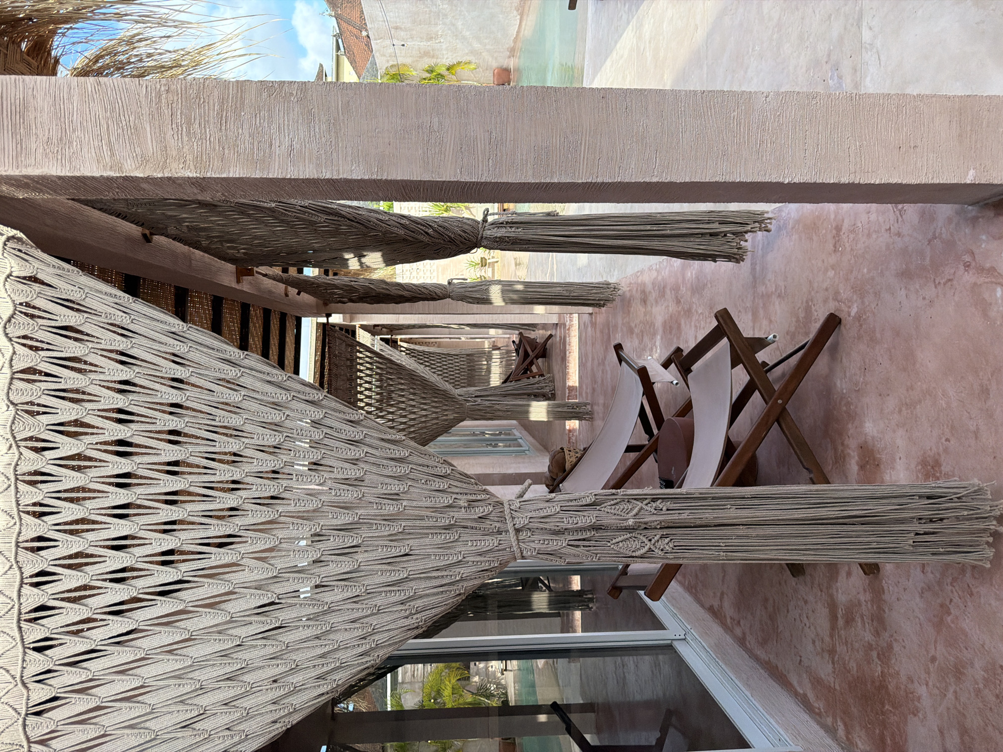 The image shows a covered patio area, likely in North Kuta, Indonesia, featuring woven hammocks strung between supporting beams. The patio has a reddish-brown concrete floor, and several folding beach chairs are positioned along the edge of the structure. A building with windows is visible in the foreground, reflecting exterior elements. The beams are textured with a light-colored stucco. Above, glimpses of the sky, neighboring buildings, and greenery can be seen.