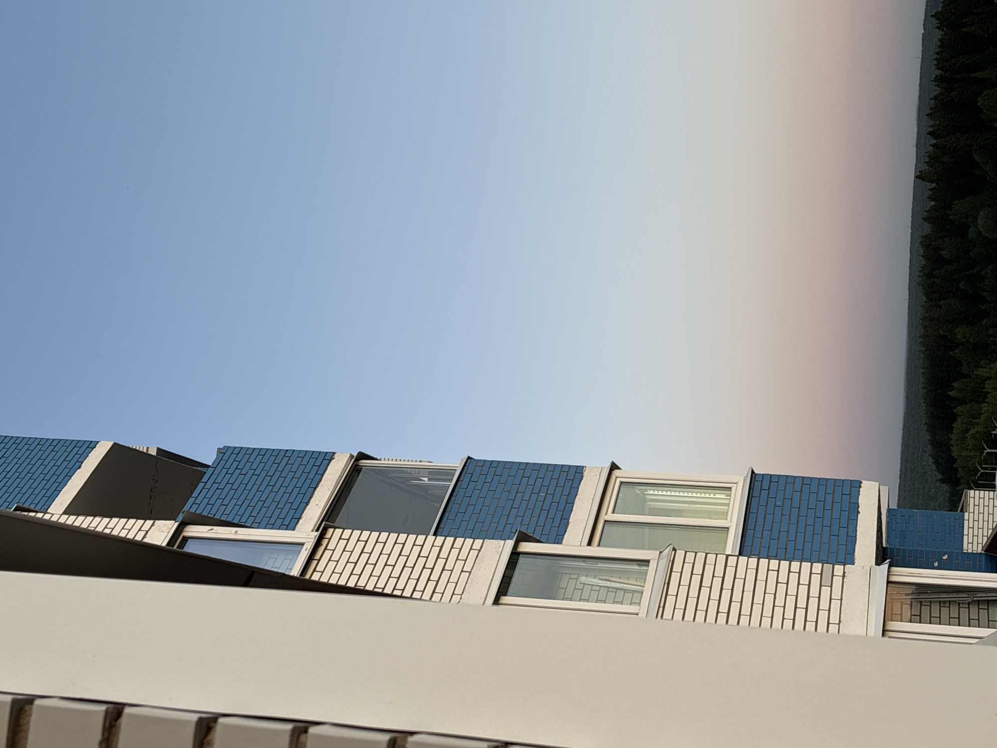 The image shows an upward angle view of a building's exterior in Izhevsk, Russia. The building has a distinctive design featuring a series of angled windows and a brick pattern that alternates between blue and white. A white wall forms the foreground. The background displays a pale sky with a gradient that suggests either twilight or a hazy atmosphere. A band of dark green, likely trees, stretches across the upper right portion of the image. The photograph appears to be taken from a low vantage point.