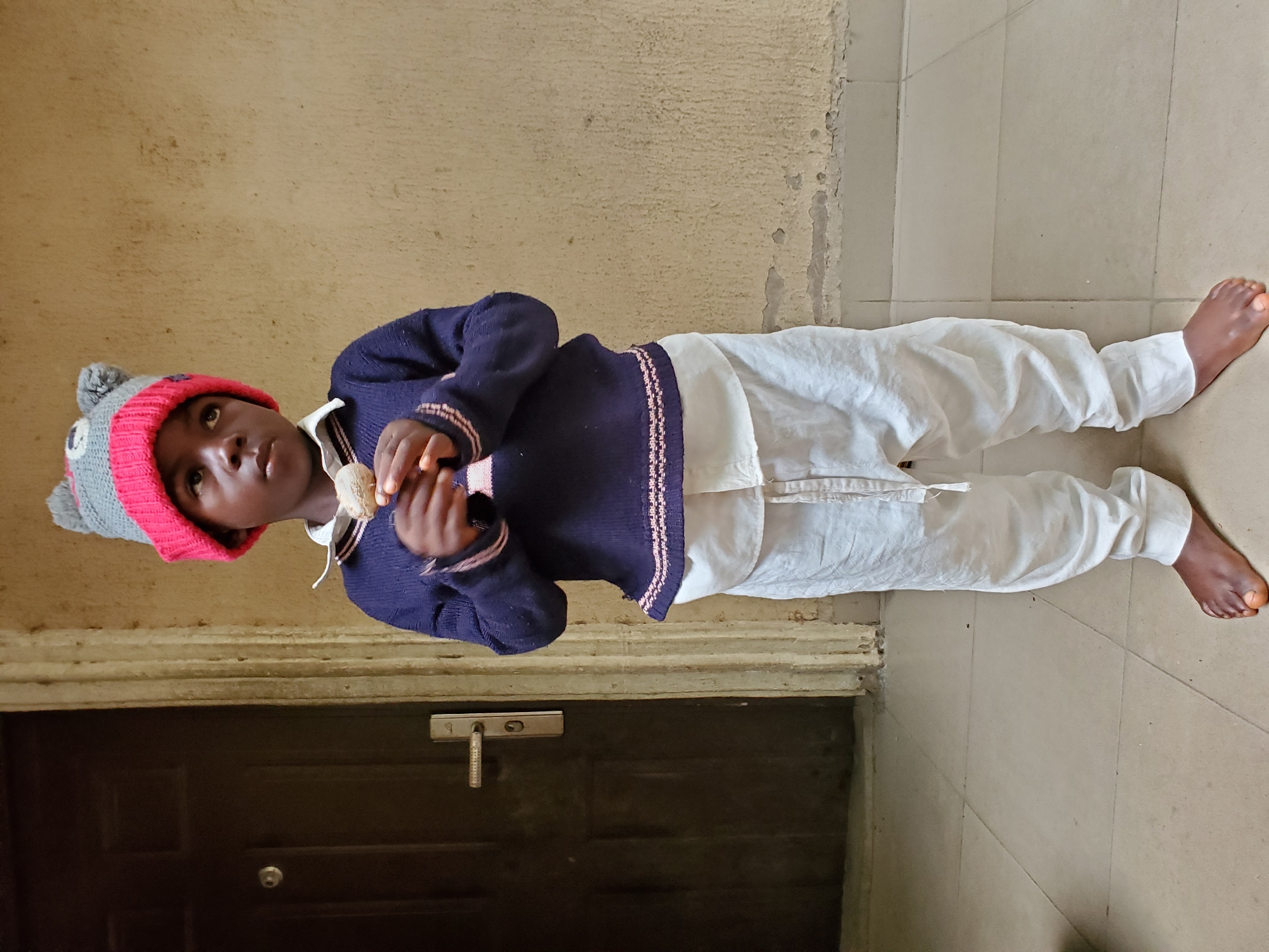 A child stands indoors in Mararaba, Nigeria. The child wears a grey and red knit hat, a navy blue sweater with pink trim, and white pants, and holds a small, round object in their hands. The background features a tan wall with visible wear and a dark brown door with silver hardware. The floor is tiled in light shades. The child is barefoot.