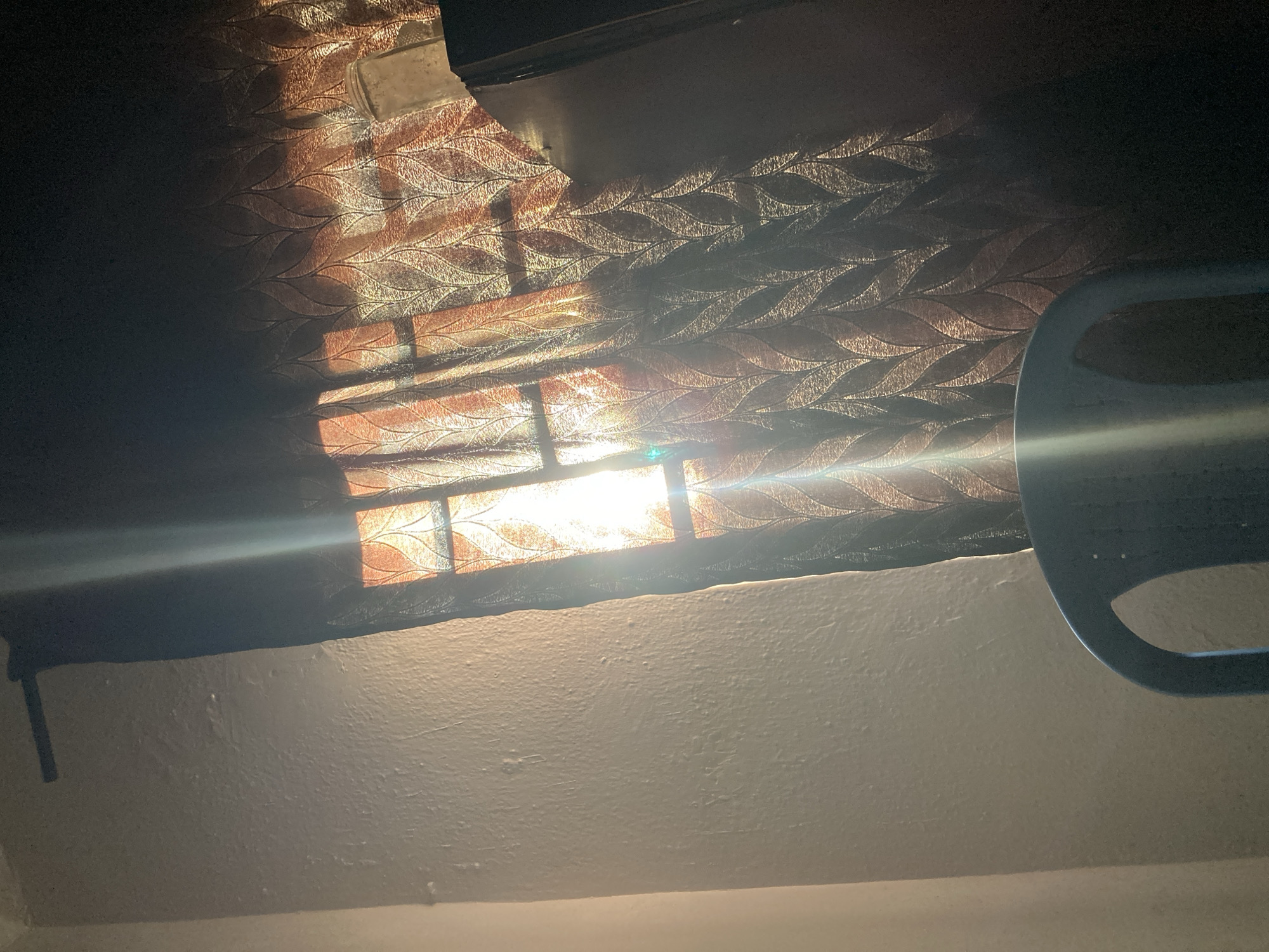 The image depicts an interior scene, likely in Jos, Nigeria, as indicated by the prompt. Sunlight streams through a window covered by patterned curtains, casting light and shadows on a textured wall. A partial view of an object, possibly a steering wheel, is visible on the right side of the frame. The room appears to be dim overall, with the sunlight creating a stark contrast.