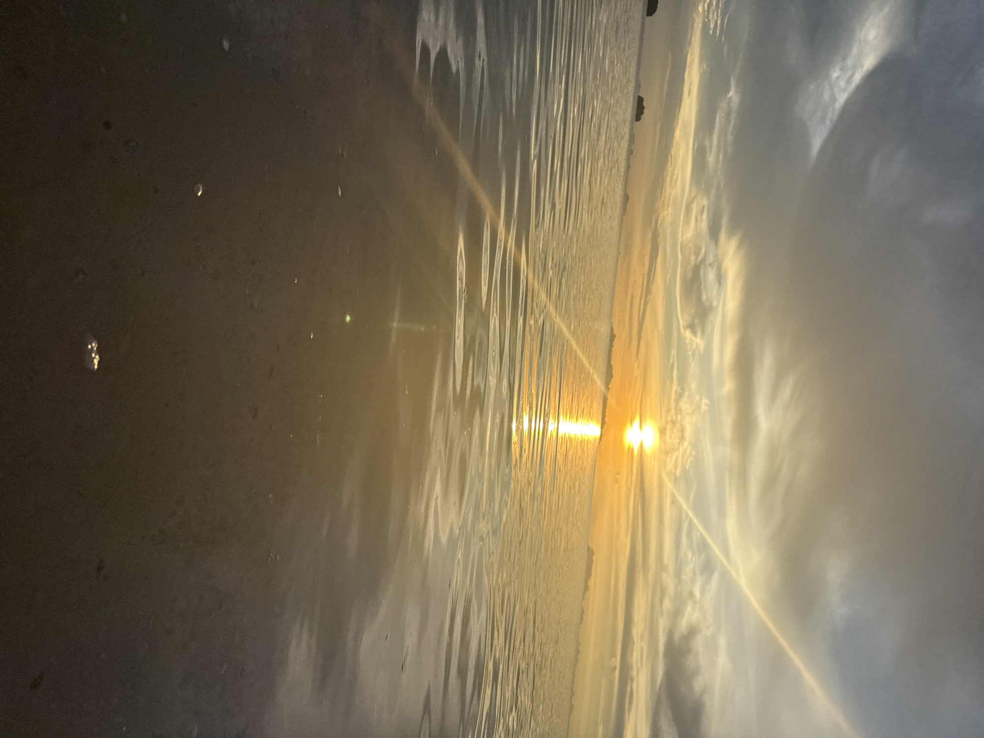 This media file captures a serene sunset over a body of water, likely the sea, given the presence of waves and a distant shoreline. The scene is dominated by the warm, golden hues of the setting sun, which casts a bright, almost blinding glare directly into the camera's lens. This intense light is reflected and refracted by the rippling surface of the water, creating a dazzling effect.
The sky is filled with dramatic clouds, ranging from soft, wispy formations to larger, darker masses that suggest a dynamic weather system. The sunlight pierces through these clouds, creating visible rays and a strong sense of atmospheric depth. A faint, almost iridescent line of light stretches diagonally across the sky and water, possibly a contrail or another atmospheric phenomenon.
On the left side of the frame, a dark, wet surface can be observed, with a few small, reflective circles that might be water droplets or small objects. This area appears to be the shore or a wet, dark ground. In the distance, on the horizon, there are faint outlines of what appear to be islands or landmasses with some vegetation.
The overall mood of the image is peaceful and awe-inspiring, typical of a beautiful sunset. The time of day is clearly evening, as indicated by the low position of the sun and the warm, golden light. The location cues, such as the tropical-looking vegetation on the distant land, along with the provided location context of Baan Pang Ka, Thailand, suggest a coastal setting in Southeast Asia. No visible text is present in the image.