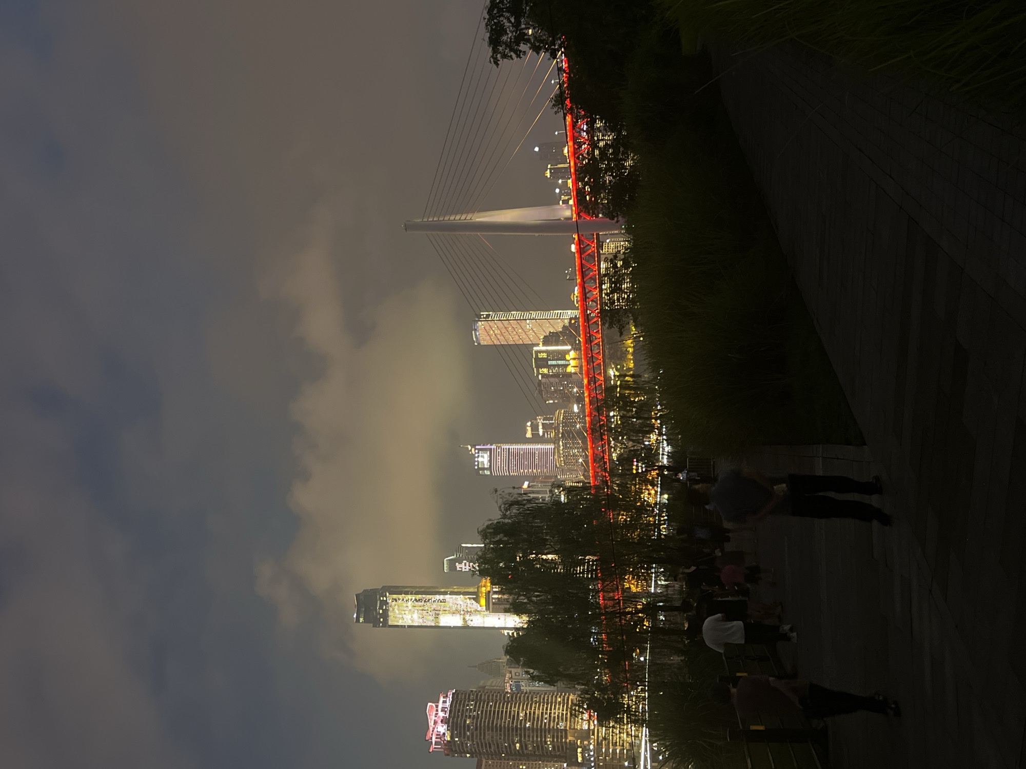 The image captures a nighttime cityscape of Jiangbei Cheng Street, China, with a focus on the illuminated skyline and a prominent bridge. The foreground features a paved walkway with several silhouetted figures of people strolling. Tall grass borders the right side of the walkway, obscuring the ground beyond. In the mid-ground, a river or body of water is visible, lined with trees and lush greenery, some of which are illuminated from behind by the city lights. A suspension bridge with bright red lighting spans across the water, connecting the two sides of the city. Behind the bridge, a dense cluster of skyscrapers, some of which are brightly lit with yellow and white lights, form the city's skyline. The sky is cloudy and dark, contributing to the nocturnal ambiance.