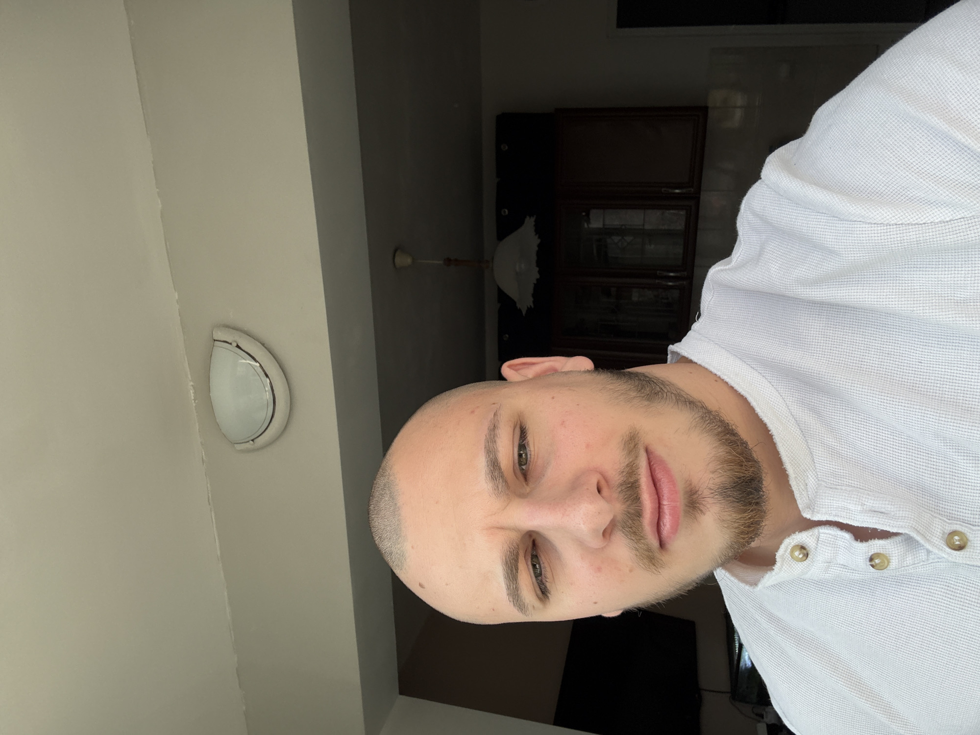 A male individual, approximately 20-30 years old, is depicted in a close-up, indoor photograph taken in Stara Zagora, Bulgaria. The subject has a very short buzz cut, light-colored eyes, and a light-brown beard and mustache. He is wearing a white, textured Henley shirt with a three-button placket, two buttons open, and tan-colored buttons. The subject directly faces the camera with a neutral expression. The background consists of a light-colored ceiling exhibiting a horizontal seam and a wall-mounted semi-circular light fixture to the viewer's left. Further back, a darker area contains a hanging pendant light with a white, bell-shaped shade and dark wood cabinets featuring glass doors, suggesting a domestic kitchen or dining setting.