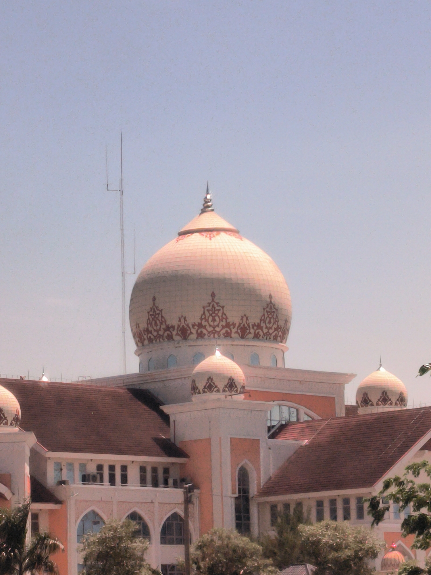 The image shows a building with multiple domes, the largest of which is a light peach color with decorative patterns near its base. The building has a combination of white and peach-colored walls and red-tiled roofs. The architecture includes arched windows and doorways. An antenna is visible in the background against a light blue sky. Trees are located in the foreground, partially obscuring the lower portion of the building. The location is identified as Meunasahaluedrienlhoksukon Barat, Indonesia.