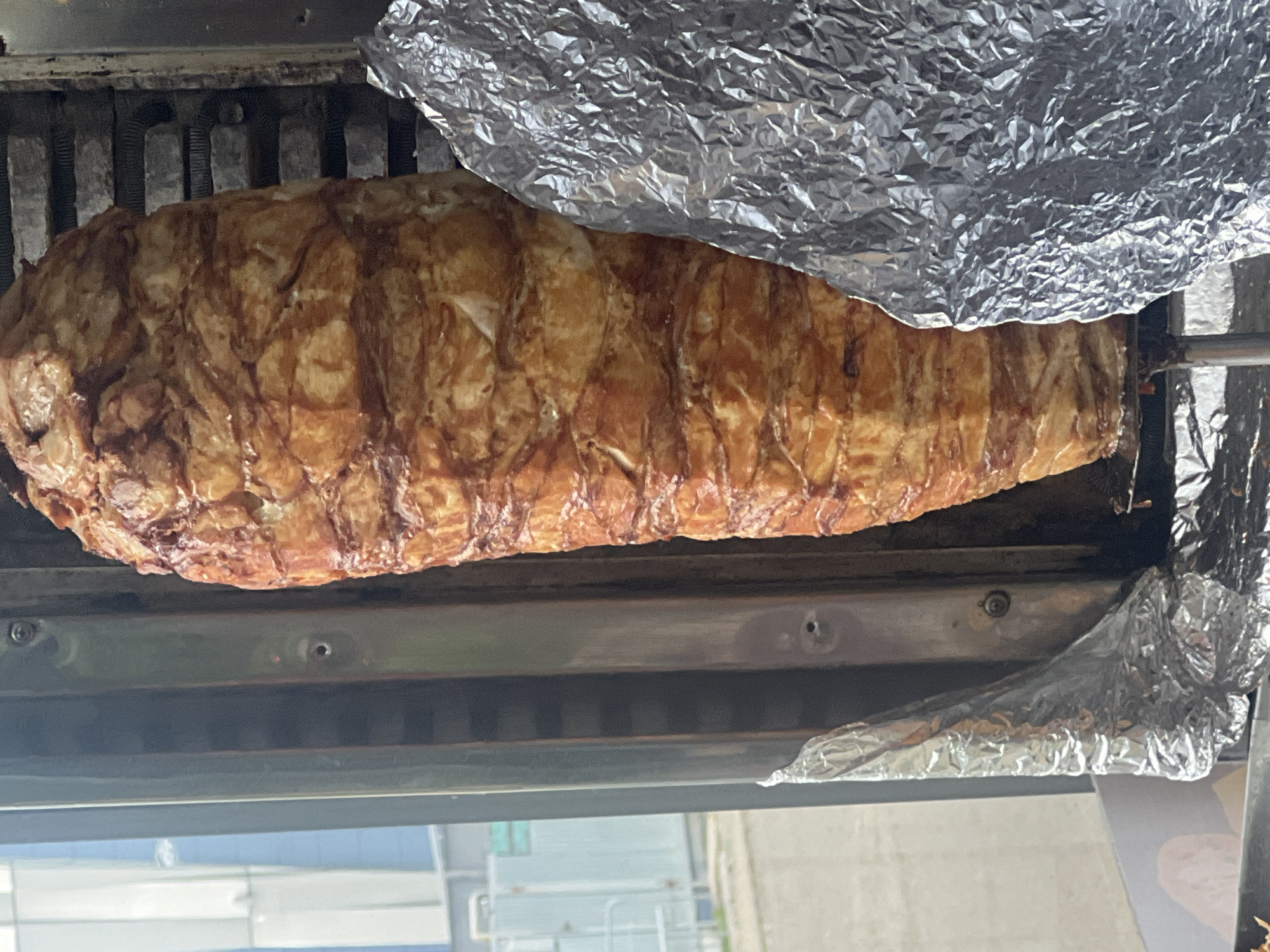 A large vertical rotisserie of seasoned meat, likely chicken or lamb, is the central subject of this close-up outdoor photograph. The meat is cooked to a golden brown with visible layers and a slightly crispy exterior. It is mounted on a spit within a metallic cooking apparatus that features heating elements in the background. To the right of the meat, a large sheet of crumpled aluminum foil is positioned, partially obscuring the cooking apparatus and perhaps serving to catch drippings or insulate. The background shows a blurry urban environment, including a modern building with blue and white panels and some metal fencing, suggesting this is a commercial food preparation area, possibly a food truck or an outdoor food stall in Chaikovsky, Russia. No people are visible in the image.