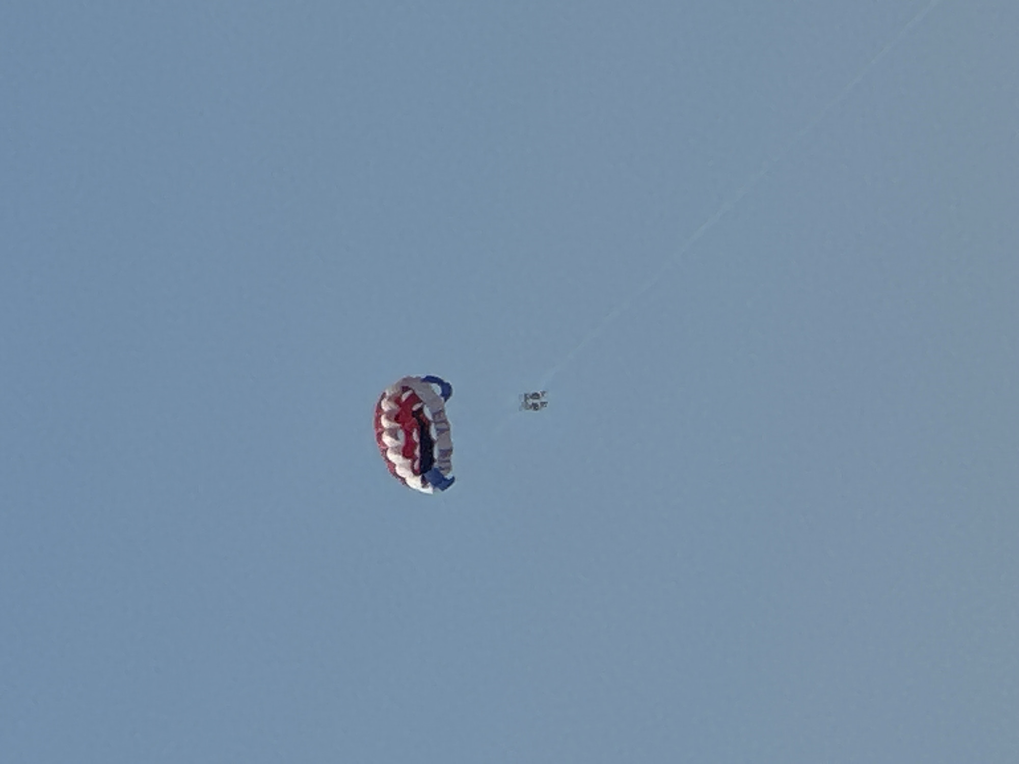 This image captures a serene sky over Budva, Montenegro, likely during daylight hours. The dominant feature is a parachute, a round one with red, white, and blue sections, suspended in the vast expanse of the pale blue sky. It appears to be in mid-descent.
A tiny, almost indistinct figure, presumably a skydiver, is visible in the upper right portion of the frame, connected to the parachute by thin lines. The skydiver is so small that details of their attire or equipment are indistinguishable. A faint, thin line, likely a tether or another string from the parachute system, extends diagonally across the upper portion of the image, further emphasizing the height and distance.
The sky is clear and unobstructed, suggesting fair weather conditions. There are no visible land features or structures, leaving the scene focused solely on the airborne objects and the immense sky. The overall impression is one of solitude and vastness, with the vibrant parachute offering a splash of color against the muted blue canvas. No text is visible in the image.