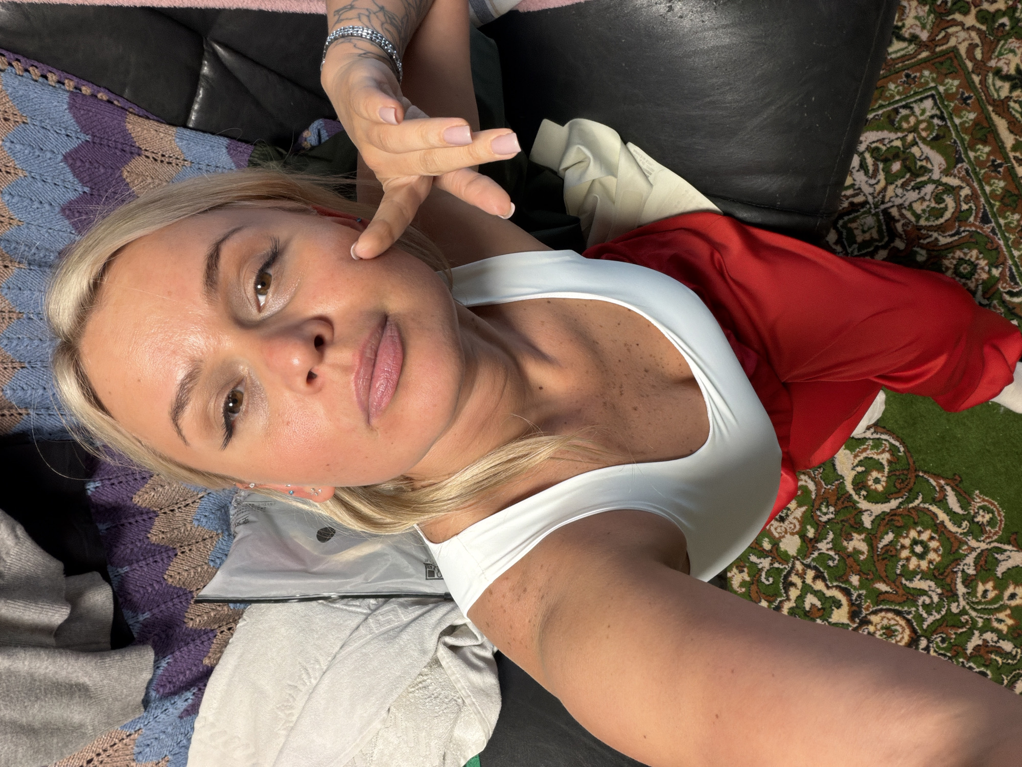 A blonde woman lies on a black leather surface. She wears a white tank top and red pants. Her left hand is near her face, and a silver bracelet adorns her wrist. A patterned blanket is beneath her, and a green carpet is visible in the background. The location in Sarapul, Russia is not discernable from this image.