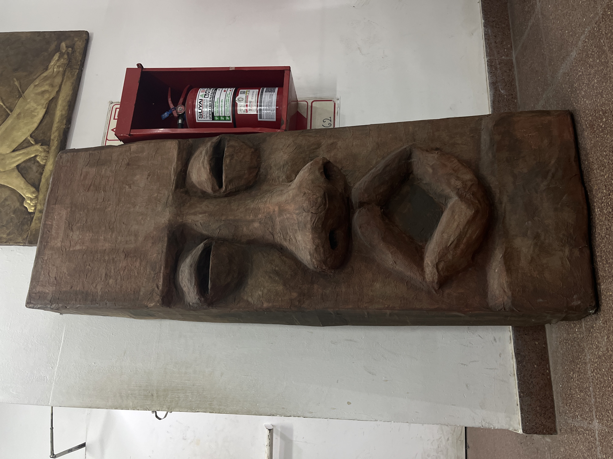This image captures an indoor scene in Buenos Aires, Argentina, featuring a large, vertically oriented art piece prominently displayed against a white wall. The artwork is a textured, dark brown rectangular panel, likely carved wood or clay, depicting a stylized human face in profile. The face exhibits a calm expression with a deep-set, elongated eye, a pronounced nose, and full, closed lips.
Above the art piece, a red metal fire extinguisher box is mounted on the white wall. Inside, a red fire extinguisher is visible, bearing the brand "SUYAI" and "EXTINTORES." Notable text on the extinguisher, in Spanish, includes "EXTINTOR A BASE DE POLVO" (powder-based extinguisher) and instructions like "COLOQUESE A 3 MTS" (place at 3 meters), along with the "A B C" fire classification. A small white sign with the number "62" is affixed to the wall just below the extinguisher box.
To the left of the main art piece, part of another golden-toned artwork, possibly a relief showing a stylized limb or wing, can be seen. The background consists of a plain white wall, and a section of speckled, reddish-brown tiled floor or lower wall is visible on the right. The setting appears to be a public building, such as a gallery, museum, or an institutional space. No human activity is depicted; the scene is static. The Spanish text on the fire extinguisher confirms the given location context of Buenos Aires, Argentina.