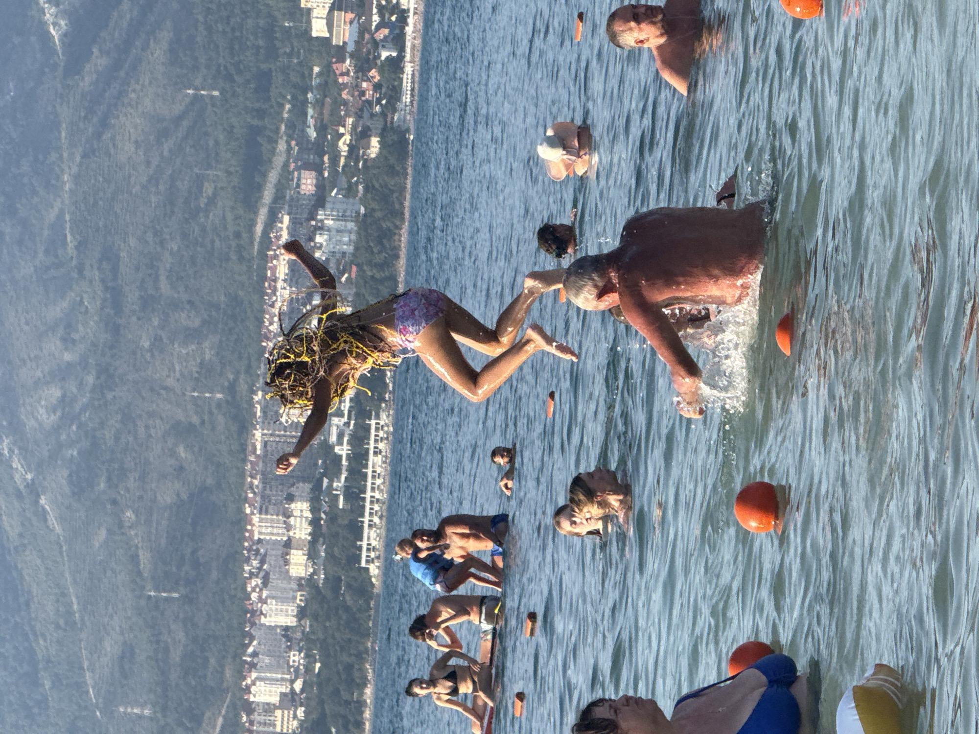 A group of people are in the sea in front of a city with many buildings and mountains behind it. The water is blue and there are some waves. In the foreground, a woman in a blue swimsuit is partially submerged in the water. To her left, an inflatable toy is floating. Several orange buoys are scattered in the water.
In the middle ground, a young woman with long dreadlocks is jumping in the air, wearing a pink and purple bikini. A man with a beard and a child on his shoulders are in the water to her left, and another man is to their right. Several other people are visible in the water, some with their heads above the surface.
In the background, a cityscape with numerous buildings is visible, nestled against a lush green mountain range. The sky is a clear blue. The overall scene depicts a lively day at the beach in Gelendzhik, Russia.