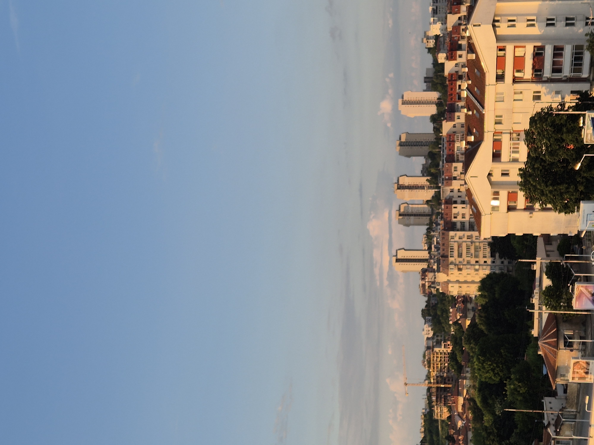 The image shows an urban landscape under a clear blue sky with a few wispy clouds. In the foreground, a white apartment building with red accents and a terracotta-tiled roof is prominently featured, bathed in the warm light of what appears to be late afternoon or early morning sun. The building has multiple stories, with numerous windows and balconies.
Behind this building, the cityscape extends, revealing a dense collection of other buildings, ranging in height and architectural style. Some are modern apartment blocks with flat roofs, while others appear to be older structures with pitched roofs. In the middle ground, a construction crane is visible, suggesting ongoing development. Further in the distance, a row of taller, light-colored high-rise buildings stands out against the horizon.
The surrounding area is characterized by lush green trees interspersed among the buildings, indicating a well-established urban environment with pockets of nature. The overall impression is a vibrant city scene, possibly captured from a vantage point on a hill, looking out over a residential and commercial district. The lighting suggests a pleasant, possibly warm, weather condition.