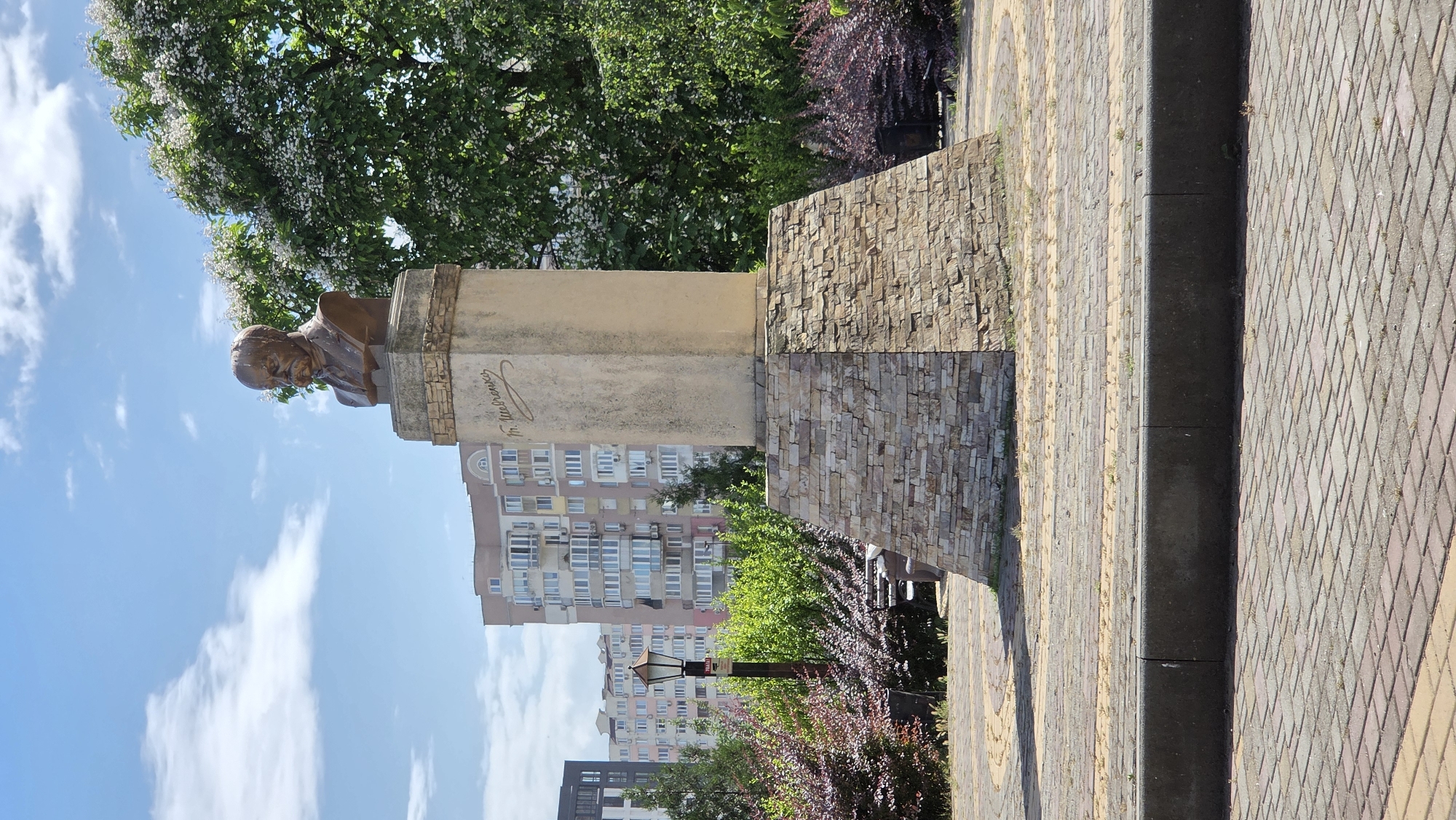 The image captures a bust of a man, presumably a historical figure given the inscription on the pedestal, which reads "Т. Шевченко" in Cyrillic. The bust is made of bronze and sits atop a stone pedestal. The pedestal itself is supported by a tiered, trapezoidal base constructed from stacked stones. The bust and pedestal are situated in an outdoor public space, likely a park or square.
In the background, modern high-rise apartment buildings are visible, suggesting an urban environment. A street lamp with a traditional design stands to the left of the monument, amidst lush green foliage and bushes with reddish-purple leaves. The ground in the foreground is paved with interlocking bricks in a pattern of yellow and gray, bordered by a darker gray curb. The sky is blue with scattered white clouds, and a large tree with white blossoms can be seen to the right of the monument.
The scene is located in the city of Pivdenne, Ukraine, as indicated by the context provided. The overall atmosphere is peaceful and sunny, with no people or animals present in the frame. The image appears to be a photograph taken from a slightly low angle, looking up at the monument.