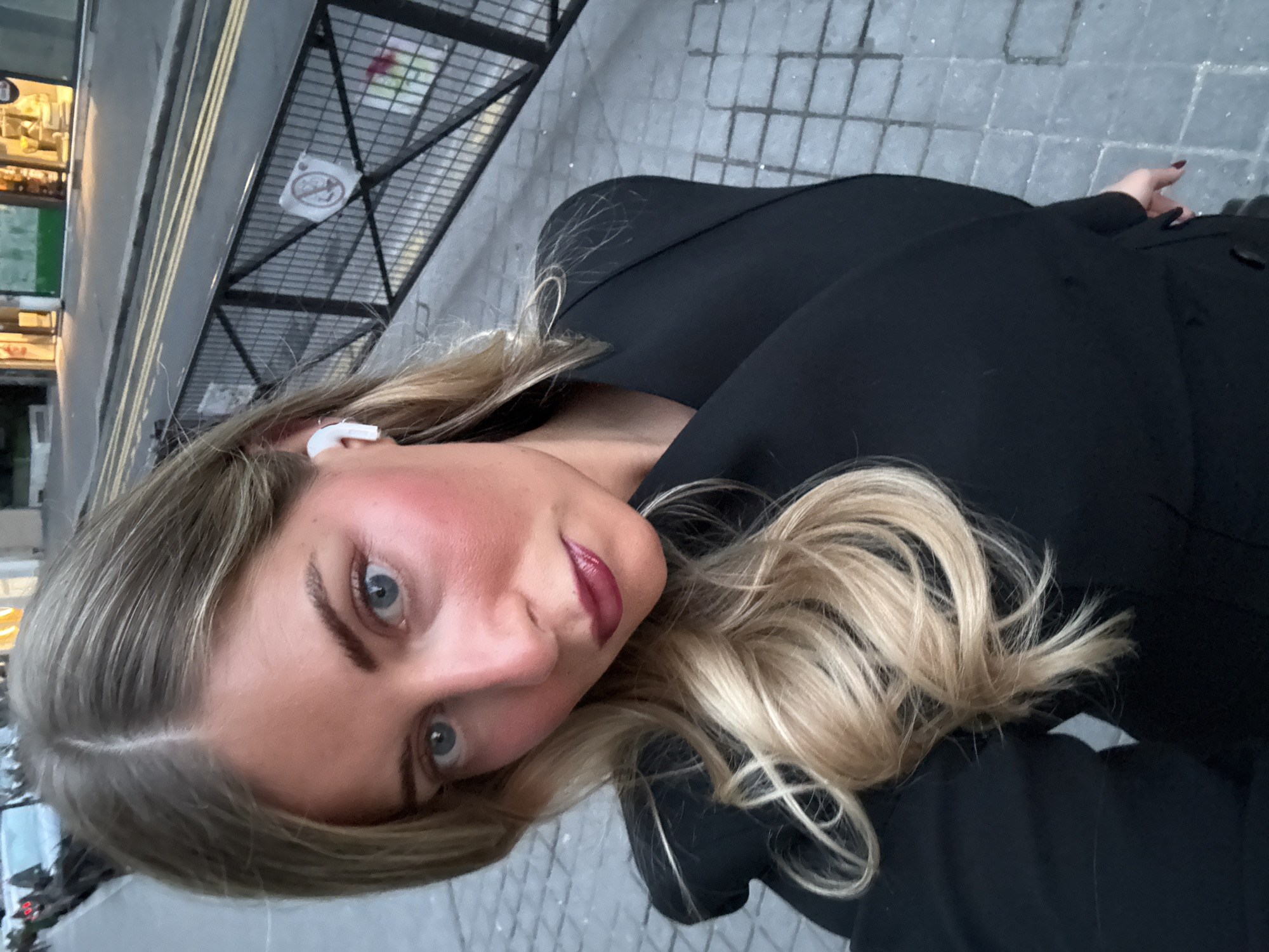 A woman with long blonde hair and blue eyes is facing the camera. She is wearing a black coat and has a white wireless earbud in her left ear. The background consists of a city street, a metal railing, a sidewalk made of grey stone, and a building. The scene appears to be in an urban environment, potentially the city of Paris, France, given the European architectural style of the building and sidewalk.