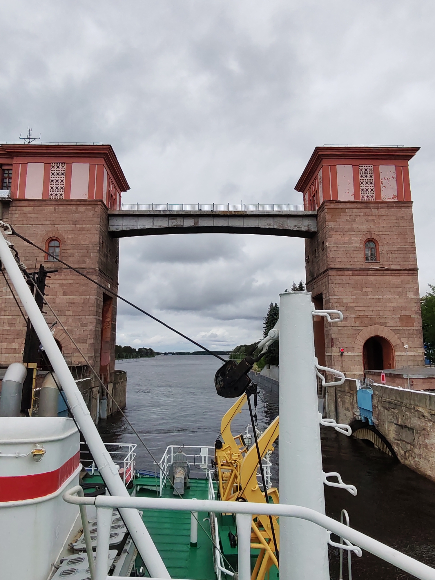 The image captures a view from the deck of a ship, looking towards a lock gate. The lock gate features two imposing towers connected by a bridge. The towers are constructed from reddish-brown stone, with red upper sections adorned with white detailing. Each tower has a single arched window. The bridge connecting the towers is made of metal. The water between the towers is visible in the background.
The ship's deck is visible in the foreground, featuring a green floor and white railings. Various pieces of nautical equipment, painted in white and yellow, are present on the deck. The sky is overcast. The location is Unknown, Unknown.