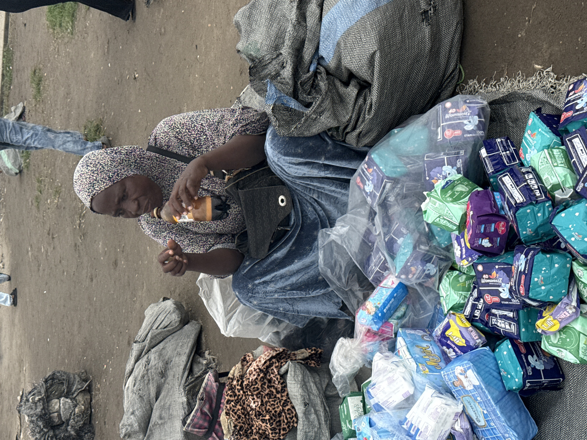 A woman wearing a headscarf and patterned dress sits on the ground in Satellite Town, Nigeria. She is holding a bottle of drink and appears to be a vendor, with stacks of sanitary pads and other goods piled around her. The ground is dusty and uneven, with other people and items visible in the background. The scene suggests a busy market or street-side selling environment.