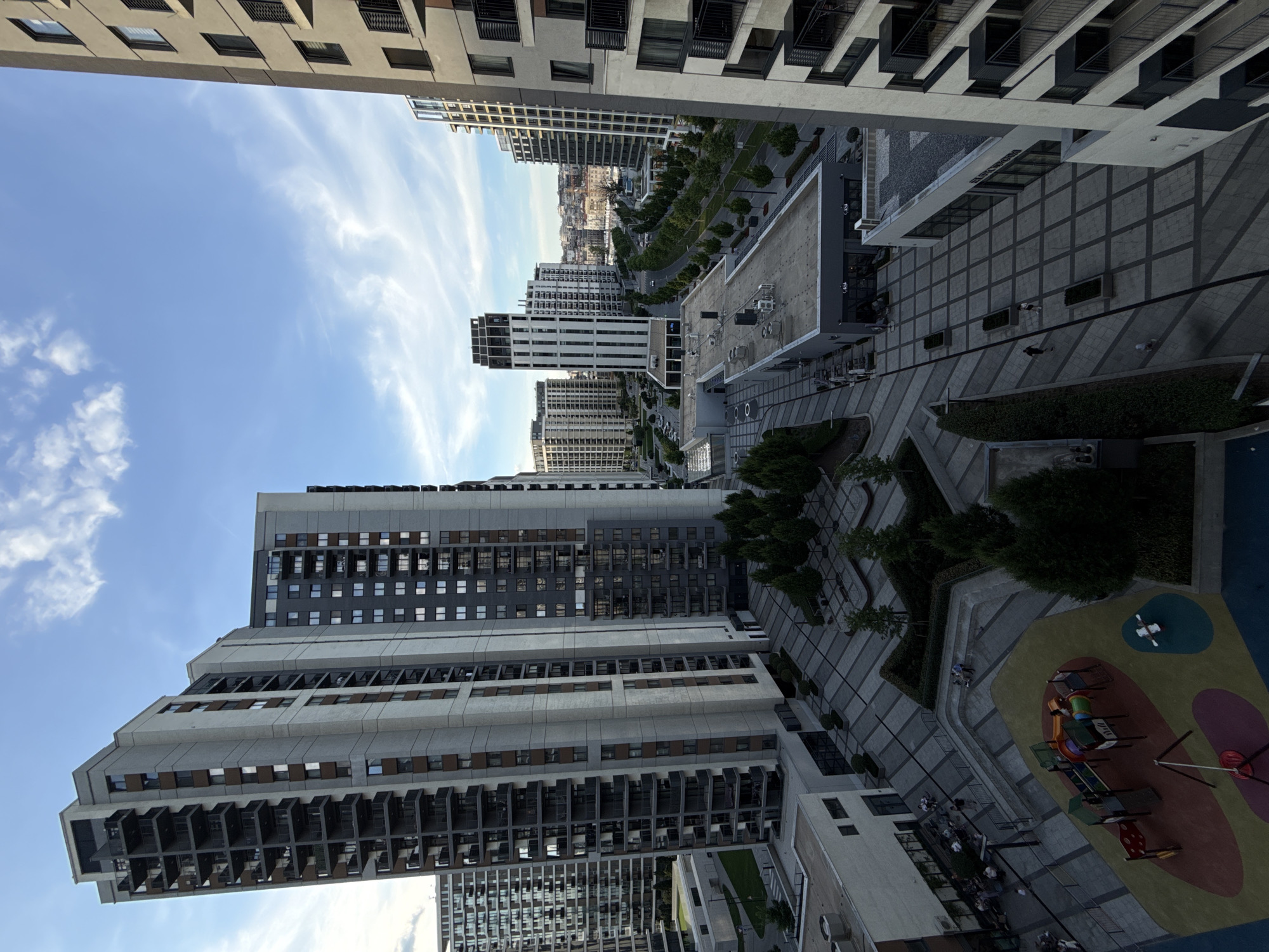 Captured from a high vantage point, the image reveals an urban setting in Unknown, Unknown, dominated by contemporary high-rise buildings. The architectural style is modern, with many structures featuring a combination of light-colored concrete and dark-framed windows or balconies. The buildings vary in height and design, adding visual texture to the skyline.
A network of paved pathways and landscaped areas occupies the space between the buildings. These areas include rows of trees, manicured shrubbery, and patches of grass, indicating efforts to integrate greenery into the urban landscape. There is a visible playground area with colorful surfaces and play equipment, suggesting a family-friendly environment.
There are people present in the scene, though they appear small due to the distance of the shot. Some individuals can be seen walking along the pathways, while others are in the playground area, possibly engaged in recreational activities.
The sky is partly cloudy, with streaks of white clouds against a blue background, which illuminates the scene with natural light.