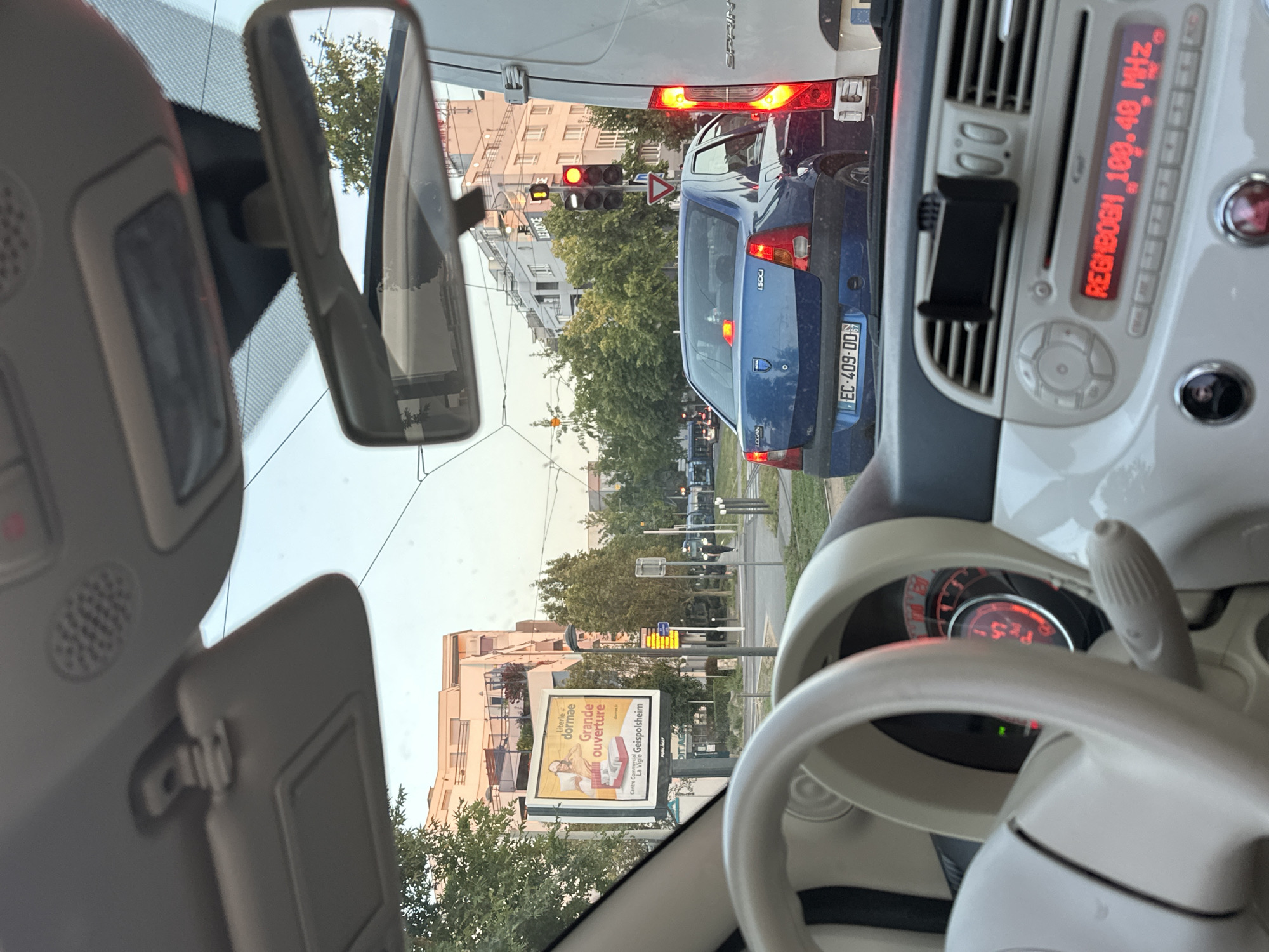 The image is a first-person perspective from inside a white Fiat 500, stopped in traffic in Strasbourg, France. The dashboard and steering wheel are visible in the foreground. Through the windshield, a blue Dacia Logan sedan is in front, with its license plate reading "EC-409-DD". To the right, the back of a white van is visible. Traffic lights are red, and a yield sign is present. A billboard in the distance advertises "Literie et dormae Grande ouverture" with an image of a man and a bed. Trees and multi-story buildings line the street. The sky is overcast and light.