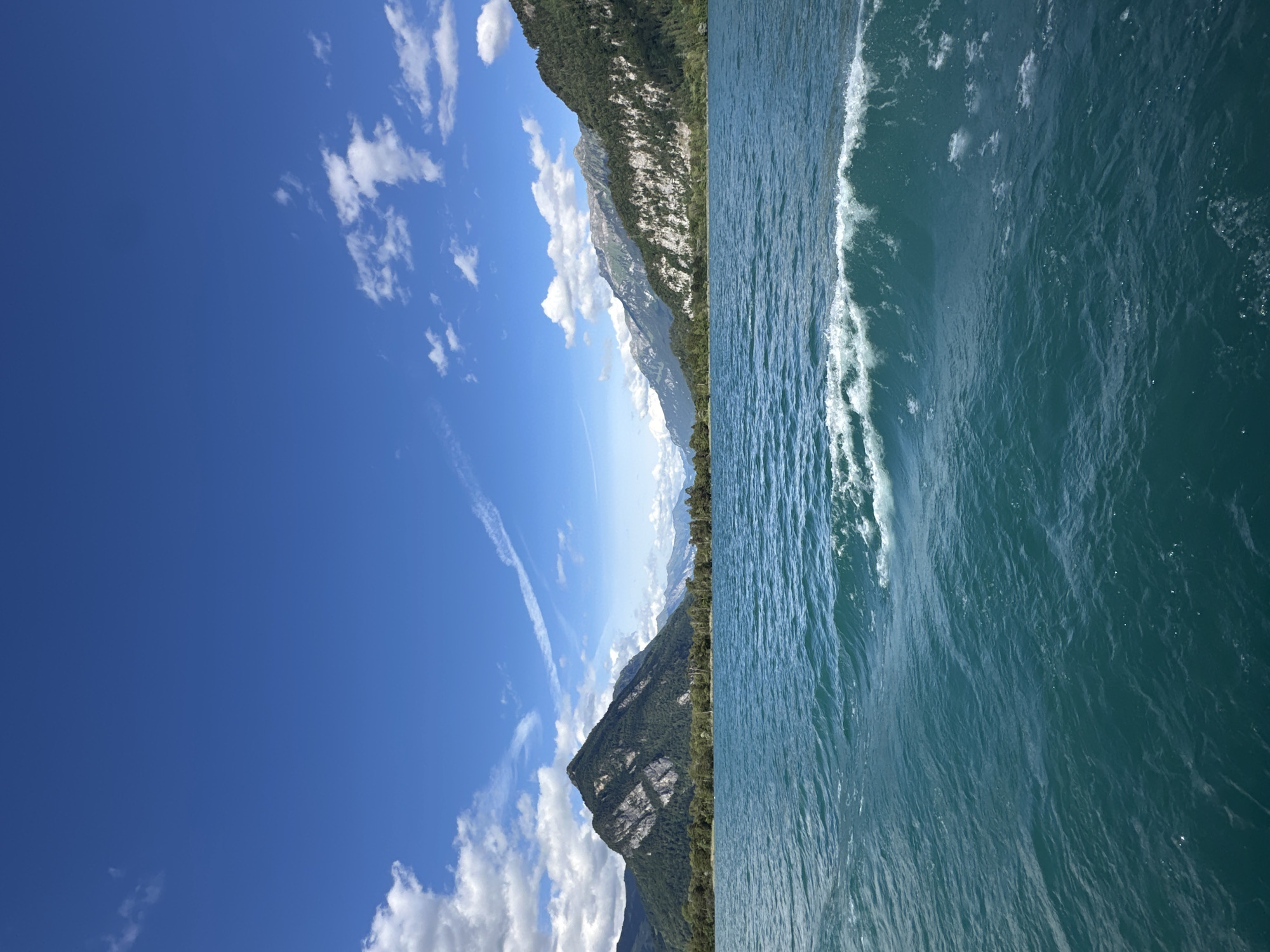 An eye-level, medium shot captures a wide, turquoise lake with a white-capped wave in the foreground. In the background, a densely wooded mountainside curves around the lake under a bright blue sky with scattered white clouds. The majestic mountain peaks are visible in the distance, some partially obscured by mist. The scene is set in Unterseen, Switzerland. There are no people or animals in the image.