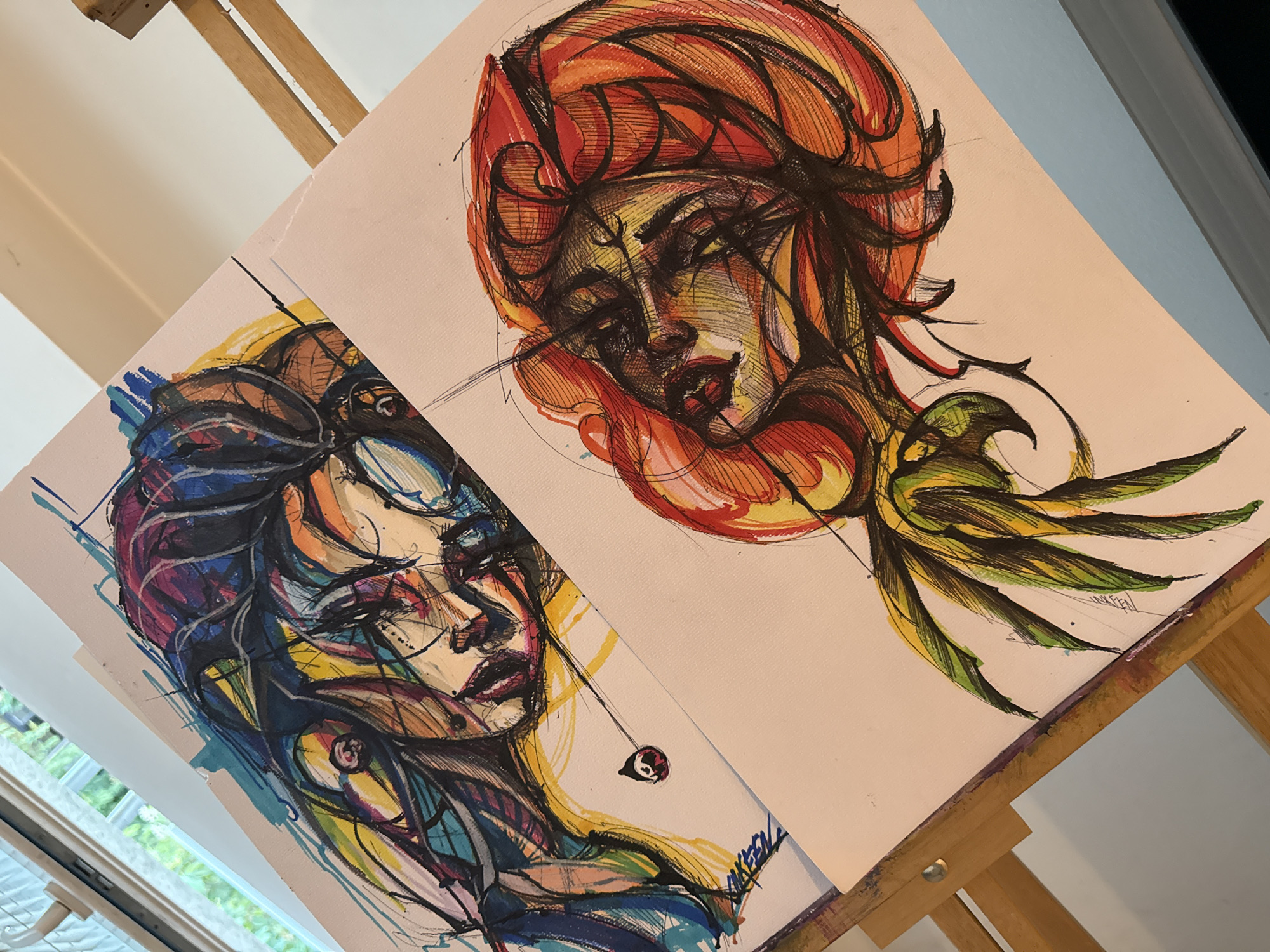 The image shows two framed drawings propped on a wooden easel. The drawing on the left depicts a woman with multicolored hair and blue and yellow skin tones, with fine black lines defining her features. She wears a dark red lipstick, and a small charm dangles near her neck. The drawing on the right shows another woman with orange and red hair, and similar fine black line work. She has a small mark on her forehead and also wears dark red lipstick. Attached to her face is a green and yellow bird-like figure. Both drawings are signed in the bottom right corner. The easel is positioned near a window with green foliage visible outside. The location in Sofia, Bulgaria cannot be determined from the image.