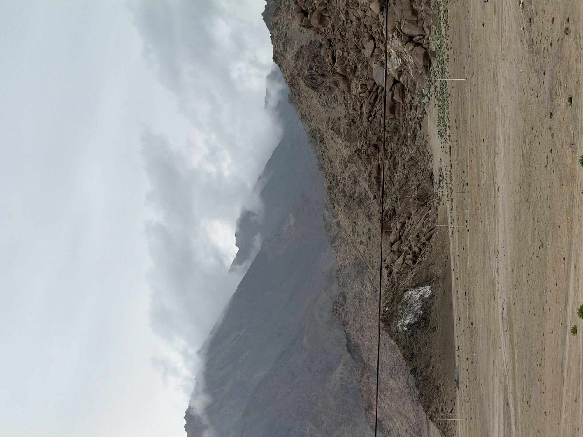 A horizontal, medium shot captures a barren, dusty landscape at the base of imposing, rocky mountains under a cloudy sky in Chumkar, India. The foreground is a vast expanse of tan-colored dirt, sparsely dotted with scrubby green vegetation. Several utility poles, thin and white, stand scattered across the plains, with dark electrical wires stretched taut between some of them. The middle ground is a steep incline of rough, brown rocks and boulders, some appearing quite large. A prominent feature in this section is a large, irregular white patch on a dark rock face. In the background, the mountains rise dramatically, their peaks partially obscured by swirling grey clouds that hug their slopes. The overall impression is one of a remote, arid, and somewhat desolate environment.