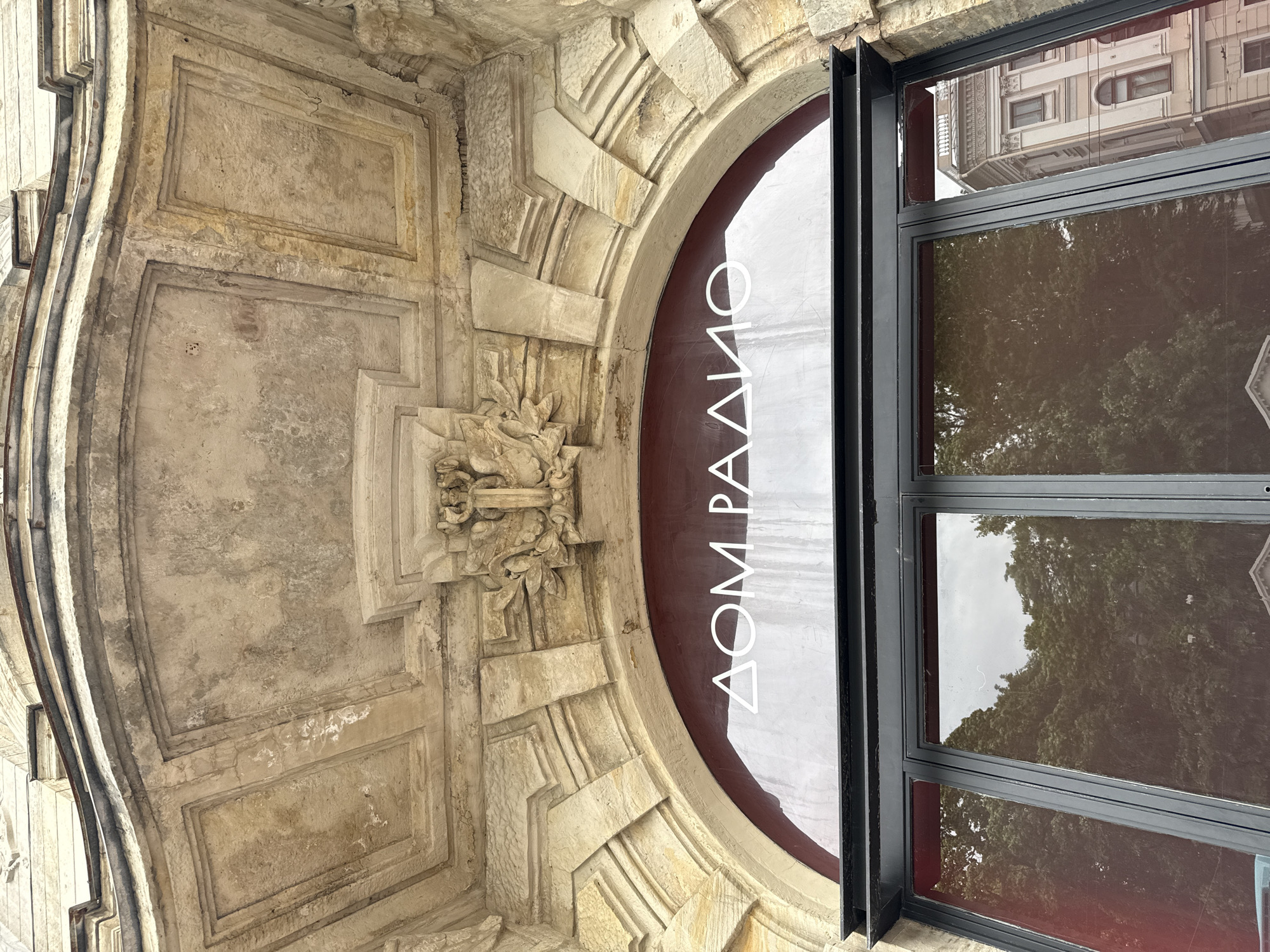 The image shows the entrance to "DOM RADIO" (House of Radio) in Saint Petersburg, Russia. The entrance features an arched glass doorway with the building's name in Cyrillic letters visible in white. Above the doorway, there is ornate stone architectural detailing, including carved decorative elements and layered stonework. The stone appears weathered and aged. The glass doors reflect trees and a building across the street. The overall scene depicts a historic facade of a cultural institution.