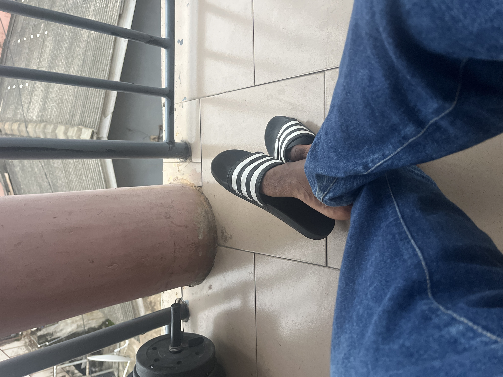A person's legs and feet, clad in blue denim jeans and black Adidas slide sandals with white stripes, are visible in the foreground of the image. The person is seated on a tiled patio or balcony, with their legs crossed. In the background, a large, reddish-brown cylindrical column stands prominently, casting a shadow. Beyond the column, a dark metal railing separates the balcony from a view of rooftops. The rooftops appear to be made of grey corrugated metal, with some buildings showing signs of age and wear. The overall environment suggests an urban setting in Accra, Ghana, possibly on a residential building's balcony or terrace. A portion of a weightlifting barbell with black plates is partially visible on the left side of the frame.