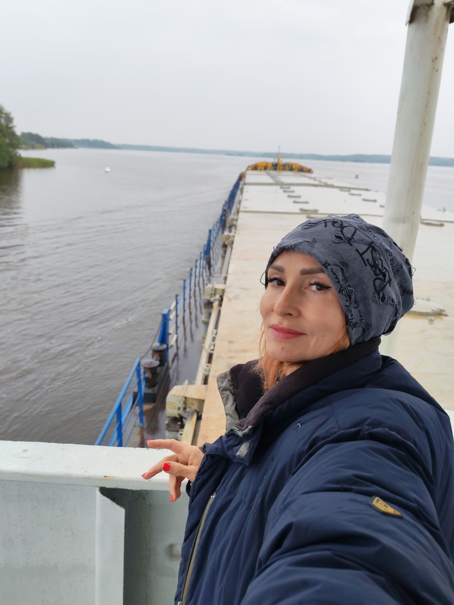 A woman with orange hair, wearing a navy blue puffer jacket and a grey beanie, stands on a ferry in Goritsy, Russia. She is looking towards the camera with a slight smile, her head tilted. Her right hand is raised, with bright red nail polish visible. The ferry is on a calm body of water, with a distant shoreline and a hazy sky. The background shows the length of the ferry, with yellow structures at the far end.