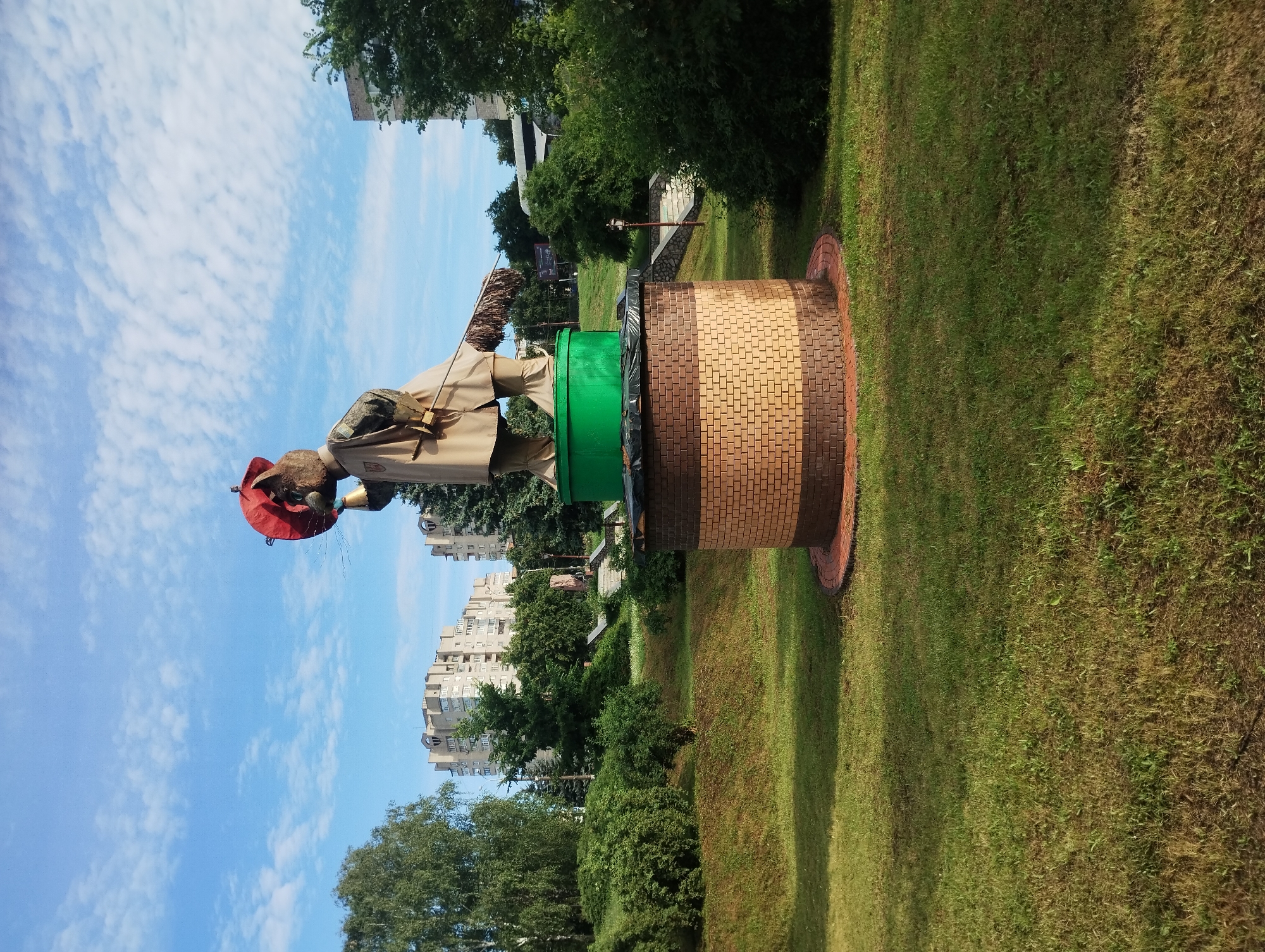A statue of a cat dressed as a musketeer stands on a circular, brick pedestal in a park in Sumy, Ukraine. The cat wears a red hat, a beige coat, and holds a red umbrella in its right paw. A broom is slung over its left shoulder. The statue is positioned in front of a green cylindrical base, which sits atop the brick pedestal. The pedestal is adorned with a pattern of light and dark brown bricks. Surrounding the statue is a well-maintained grassy area with a gentle slope. In the background, there are trees, bushes, and apartment buildings. The sky is blue with scattered white clouds. The overall scene suggests a public park area on a bright, sunny day.
