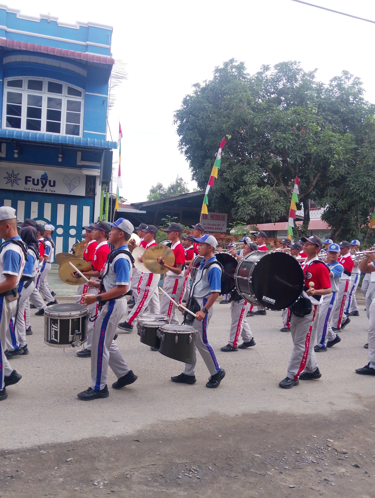 The image depicts a marching band procession on a street in Serbelawan, Indonesia. In the foreground, several young individuals in uniform are playing drums and cymbals. The uniforms consist of gray pants with blue and red stripes and white shirts with blue accents, and some wear baseball caps. In the background, a blue building with white window frames is visible, featuring a sign that reads "Fuyu Ice Cream & Tea". Further down the street, more band members are seen, some holding brass instruments. Colorful flags are displayed, adding to the festive atmosphere. The ground is paved with asphalt and shows signs of wear. The overall scene captures a community event or parade.