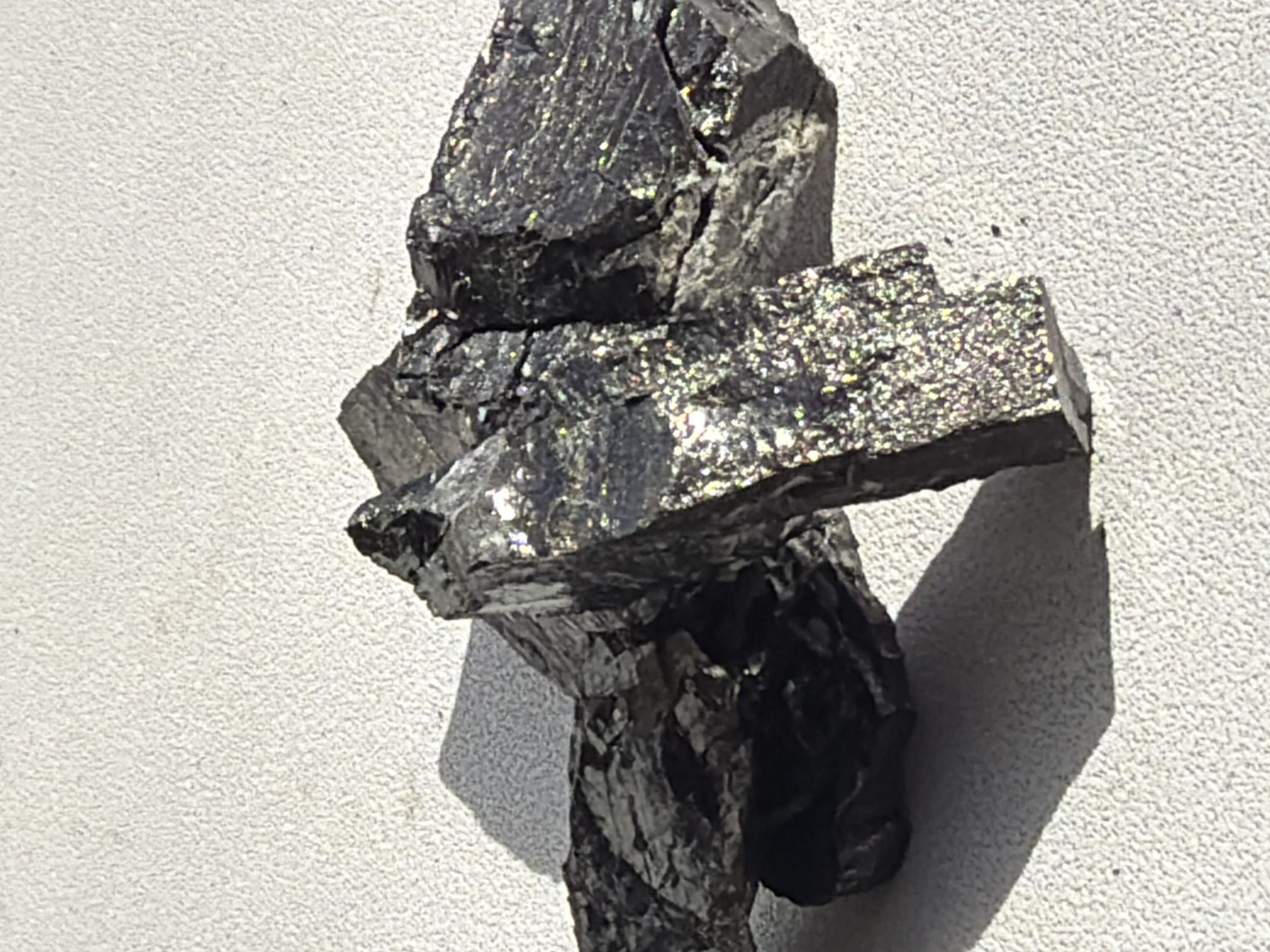 The image shows a dark, possibly metallic or mineral specimen, featuring sharp edges and an irregular surface with reflective patches. It appears to be resting on a rough, light-colored surface, possibly a wall or display platform. Strong lighting from the side casts a sharp shadow. There is no information in the image that indicates any location in Izhevsk, Russia. The image appears to depict a geological specimen rather than a specific location. There are no people, actions, or interactions visible in the image.