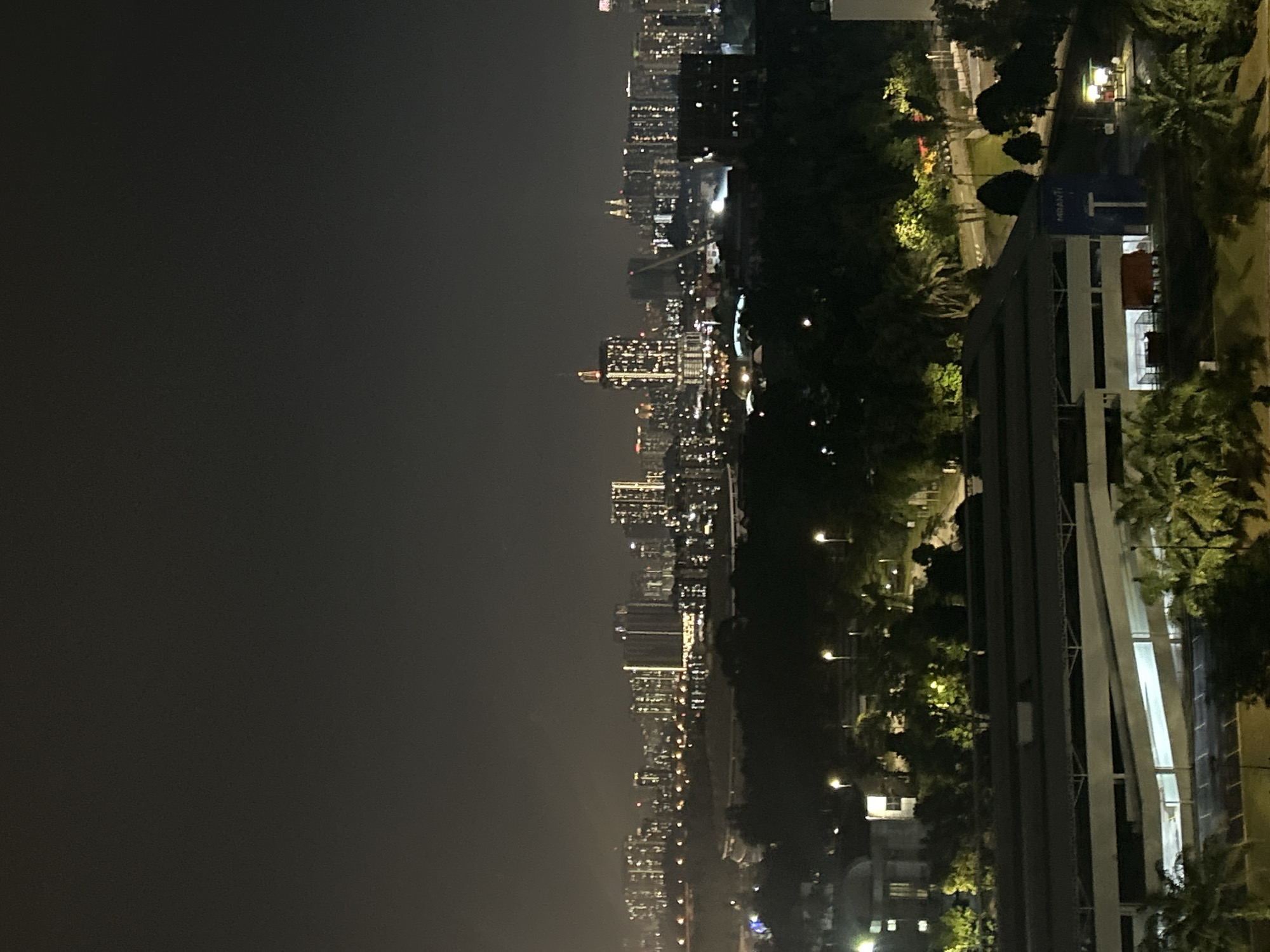 The image depicts a nighttime cityscape, likely Kuala Lumpur, Malaysia. The shot is taken from a high vantage point, possibly a building. The right side of the frame shows a modern building with a long, covered walkway or sky bridge structure extending alongside it. Trees and landscaping are visible around the building. The left side of the frame is dominated by the city skyline at night, showing numerous lit buildings extending towards the horizon. The skyline includes a dense cluster of high-rise buildings with twinkling lights indicating offices and residences. The sky above the city is dark, suggesting it is late at night.